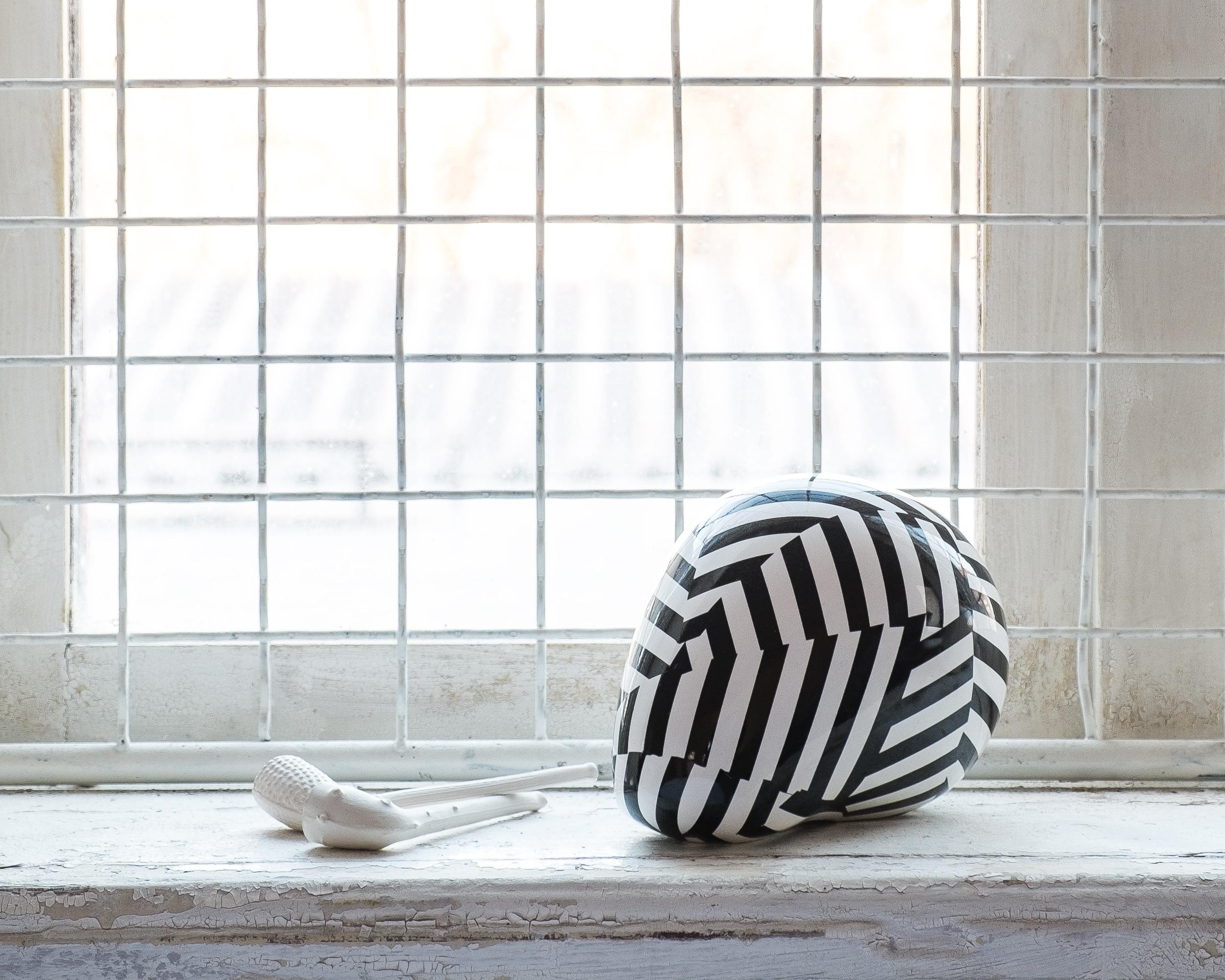 A shiny white and black porcelain figurine/sculpture titled Black Stripe is a human skull redefined as a simplistic industrial form with in-glazed imagery of dazzle camouflage hiding its true nature. Modeled in CAD, 3d printed, and then cast in