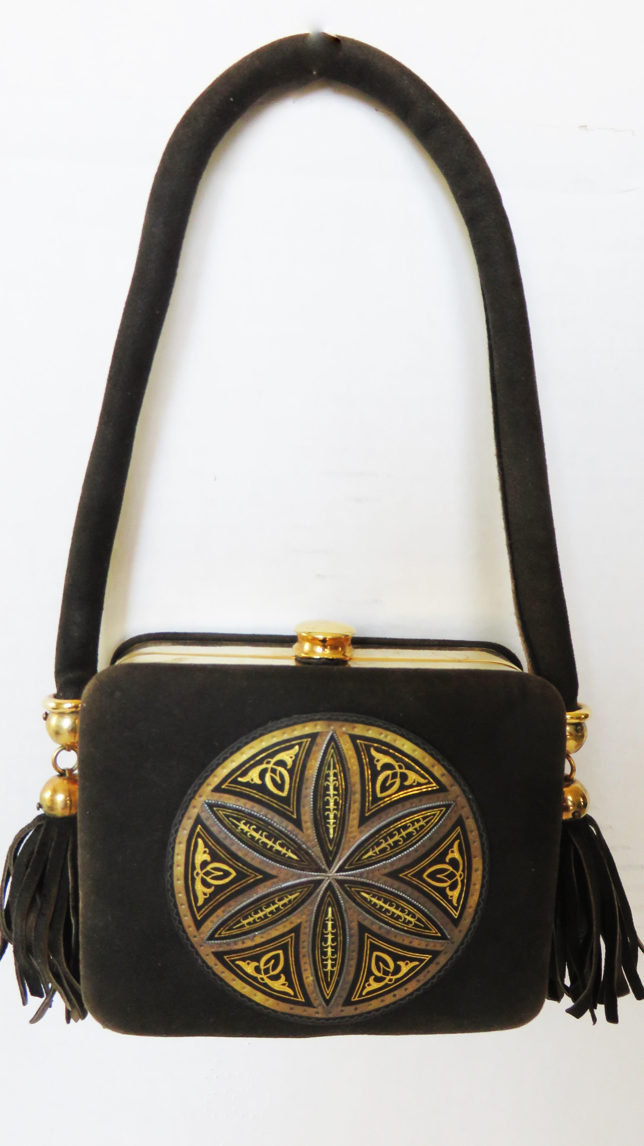 A beautiful black suede minaudiere compact wallet evening bag.  It has a top handle with suede fringe tassels on the ends, a gold metal top snap button closing and frame, and a detailed circular elaborately detailed medallion patch on the front. The