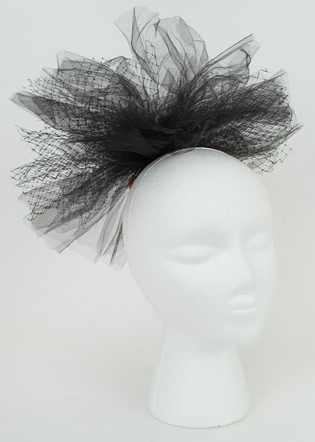 Mit der Leichtigkeit eines Fascinators und der Pracht eines Cocktailhutes formt dieser außergewöhnliche Haarschmuck eine riesige Blüte aus schwarzem Tüll und Netz. Ein Accessoire, das größer ist als der Kopf, der es trägt, und das auch als Korsage