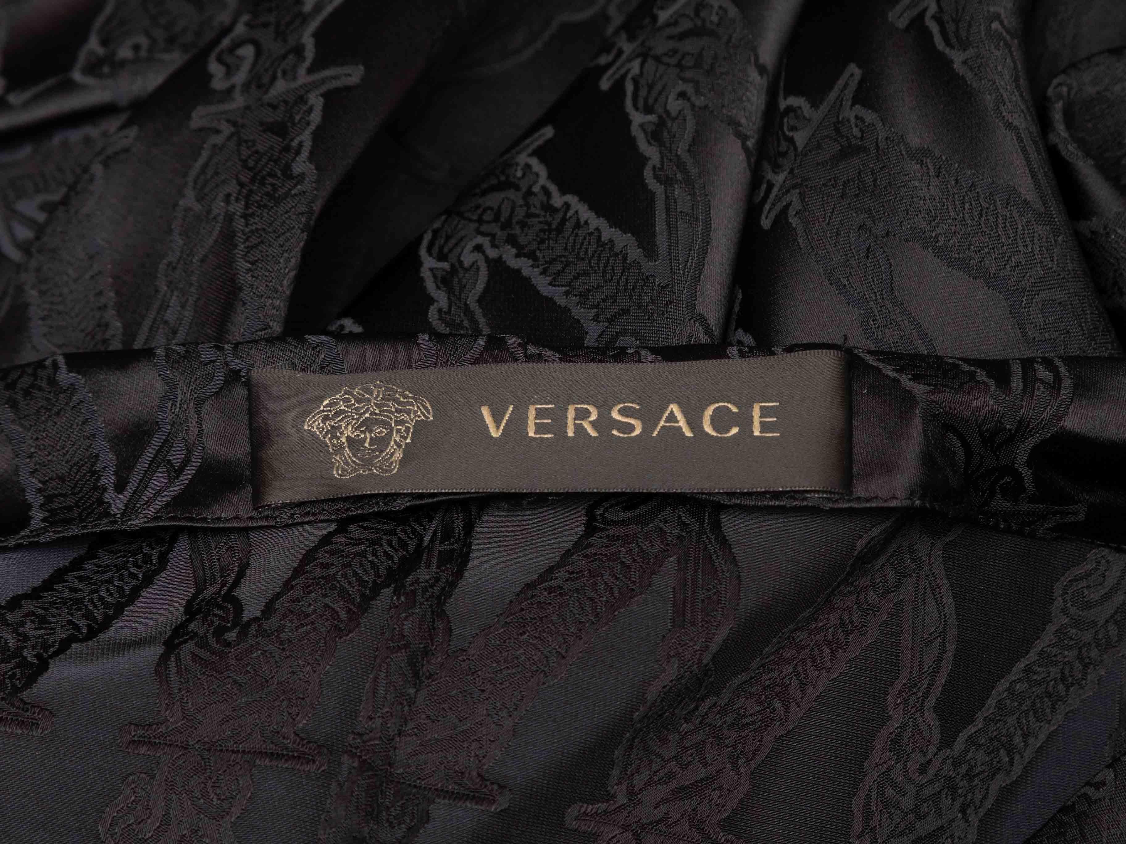 Robe à manches longues en jacquard noir, Versace. Extrait de la Collectional Automne/Hiver 2019. Encolure asymétrique. Le corsage est orné d'une chaîne en métal doré. Ourlet effiloché. Buste de 29