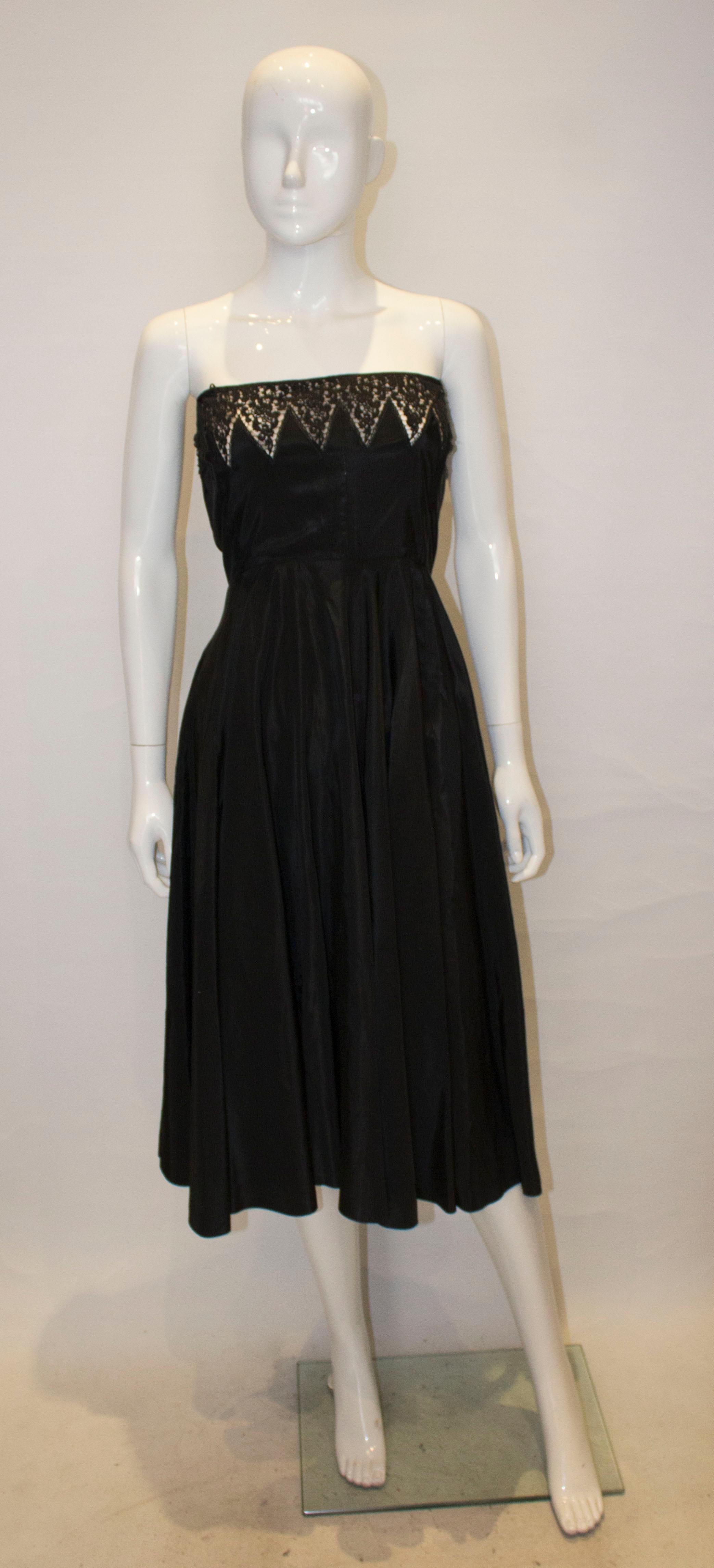 A chic black vintage cocktail dress from the 1950s., The dress is strapless, but straps could easily be added with lace detail on the bust area. It has a side zip opening and a flared skirt.