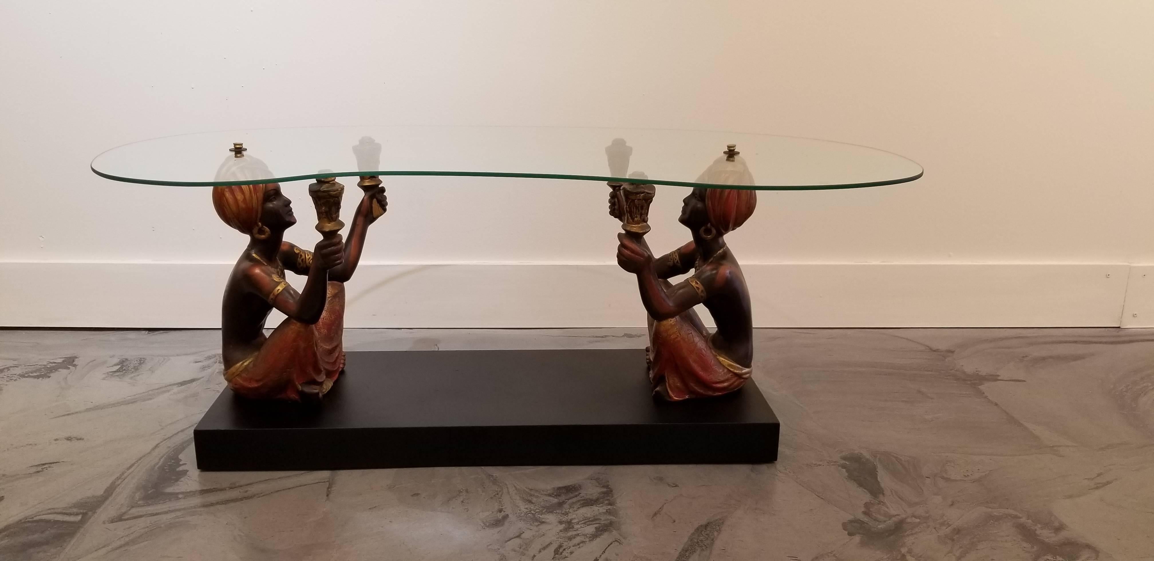Kidney shaped glass top coffee table with two polychrome Nubian figures. Original painted surface to cast plaster figures. Mounted to a painted wooden base.
