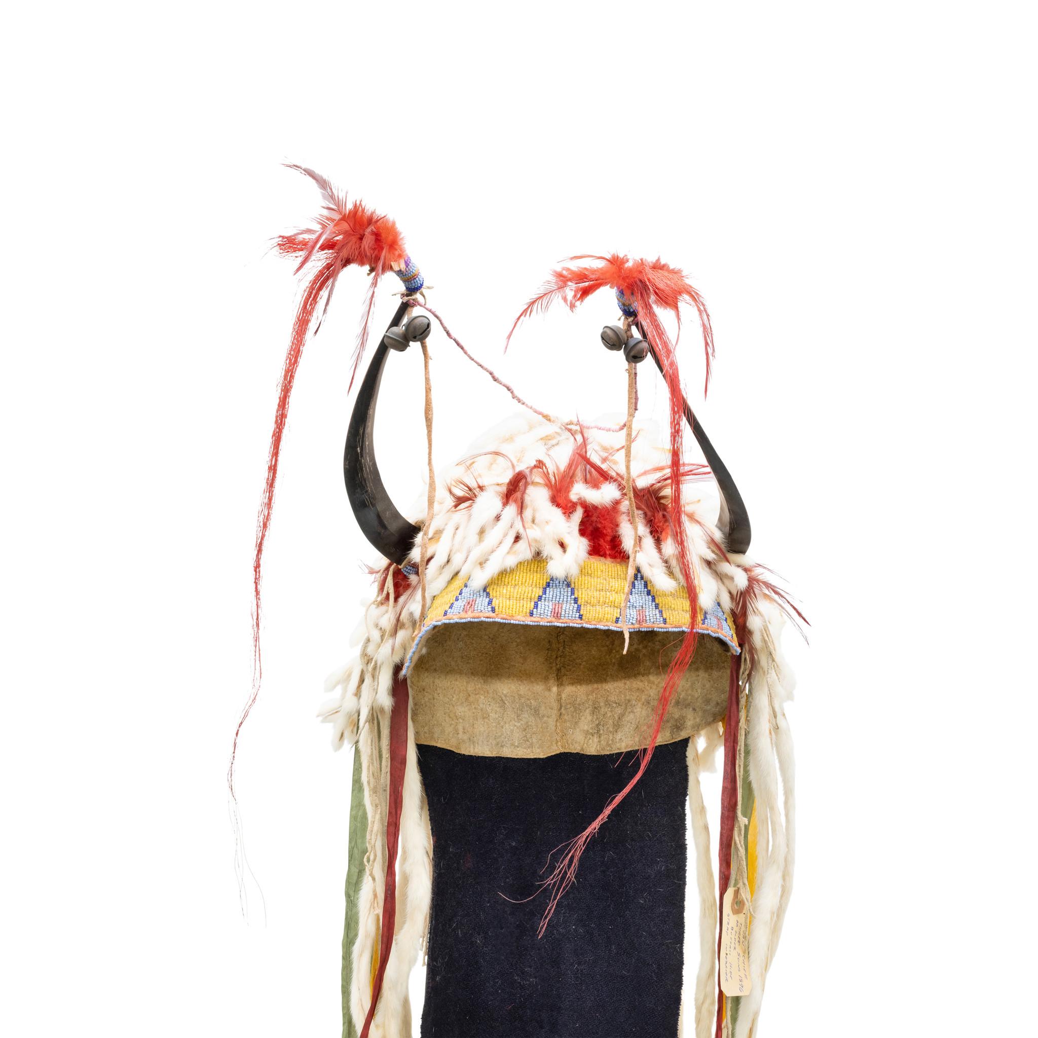 Blackfeet split horn bonnet. Sinew-sewn stroud hide cap with lazy-stitch beaded headband on brain tanned buffalo. The horns terminating in bells, beads, feather, and horsehair drops. The sides with silk ribbon drops and ermine, with blue dyed trade
