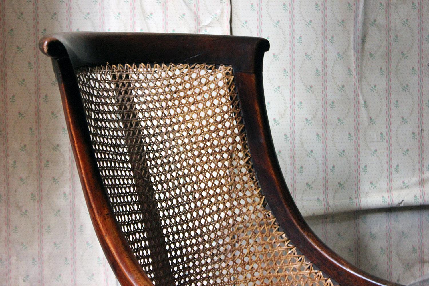 In the manner of Gillows, the Regency period mahogany framed bergère library chair having an elegant swept framed back to rear sabre legs and turned front legs, with a caned seat and back, the left flank with a large blacksmith repair of iron