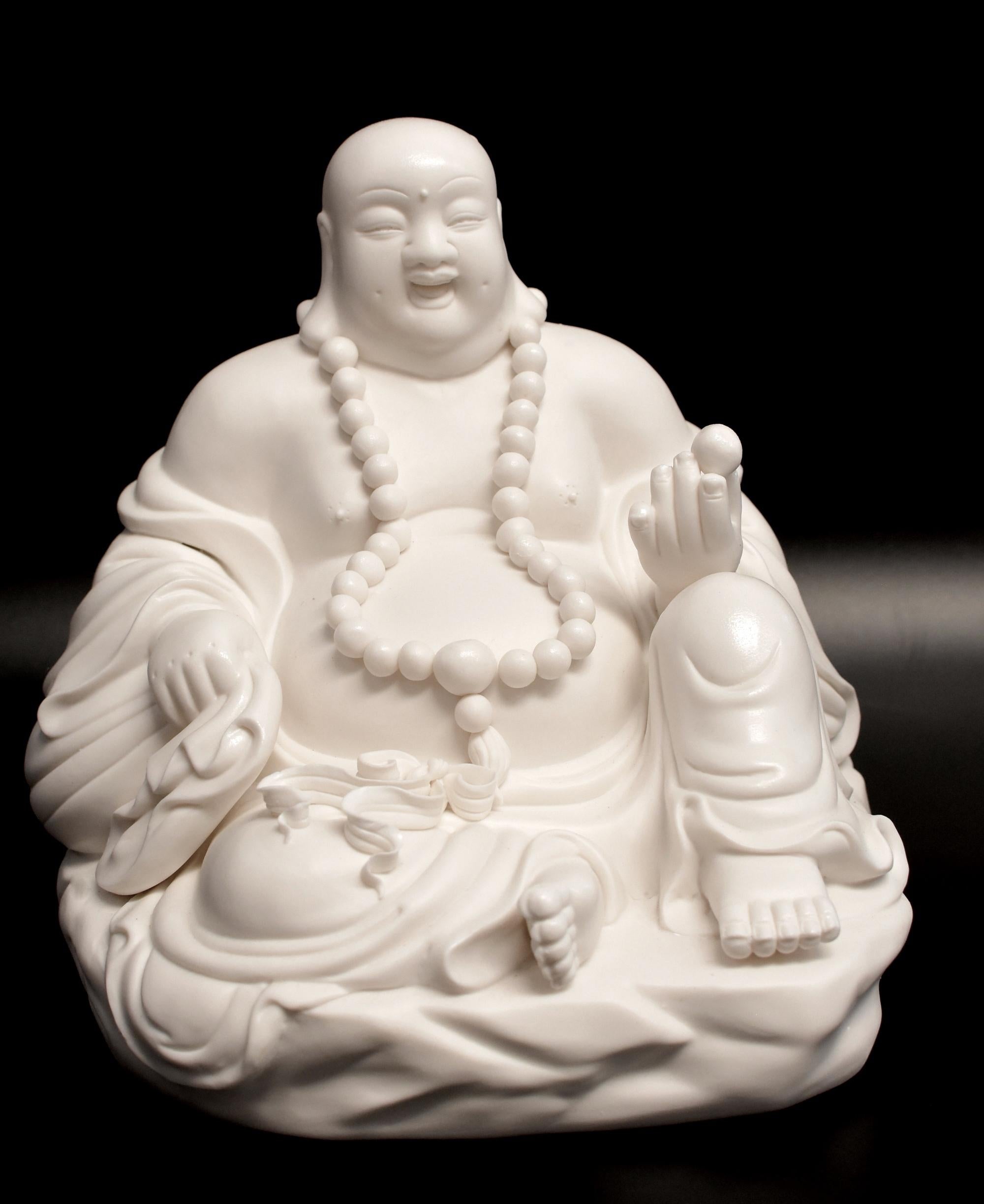 An exquisite Blanc De Chine Porcelain happy Buddha, his broad face with a joyful expression, long slender eyes beneath arched brows above an open mouth, all flanked by long pendulous ear lobes. Rope of prayer beads and a pearl in one hand symbolize