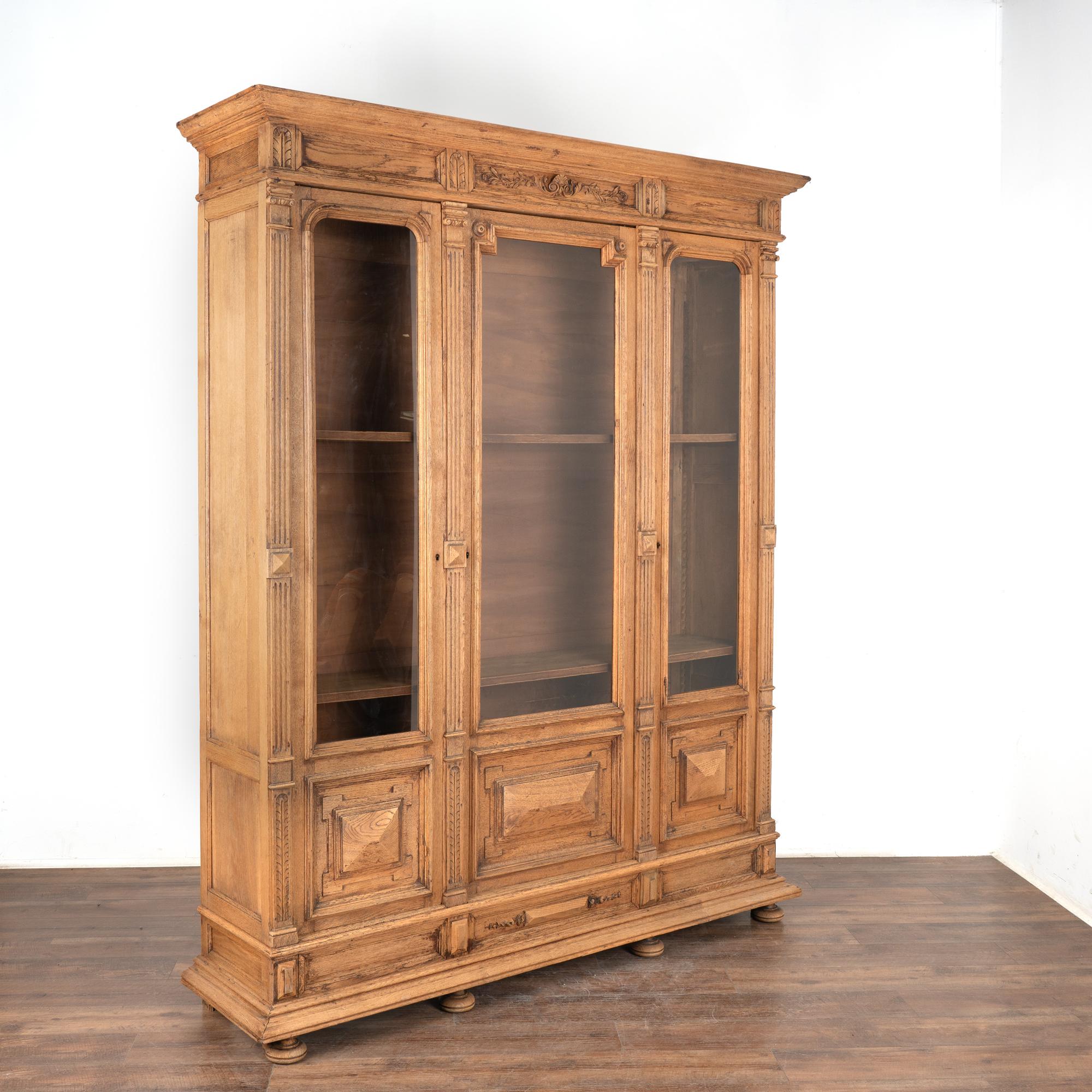 This large bleached oak bookcase or display cabinet has three long glass panels with heavy carved panel on bottom.
Ideal for display of books or any collection, the three interior shelves are adjustable (pegs included).
Notice in photos there is a