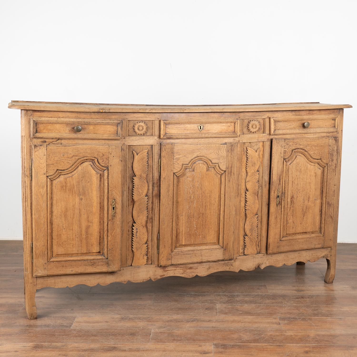 Large French bleached oak sideboard with carved accents between the three panel doors. Over 7' long, this will serve as an impressive buffet resting on carved feet.
The heavy distress along the top reflects many generations of use including cracks,