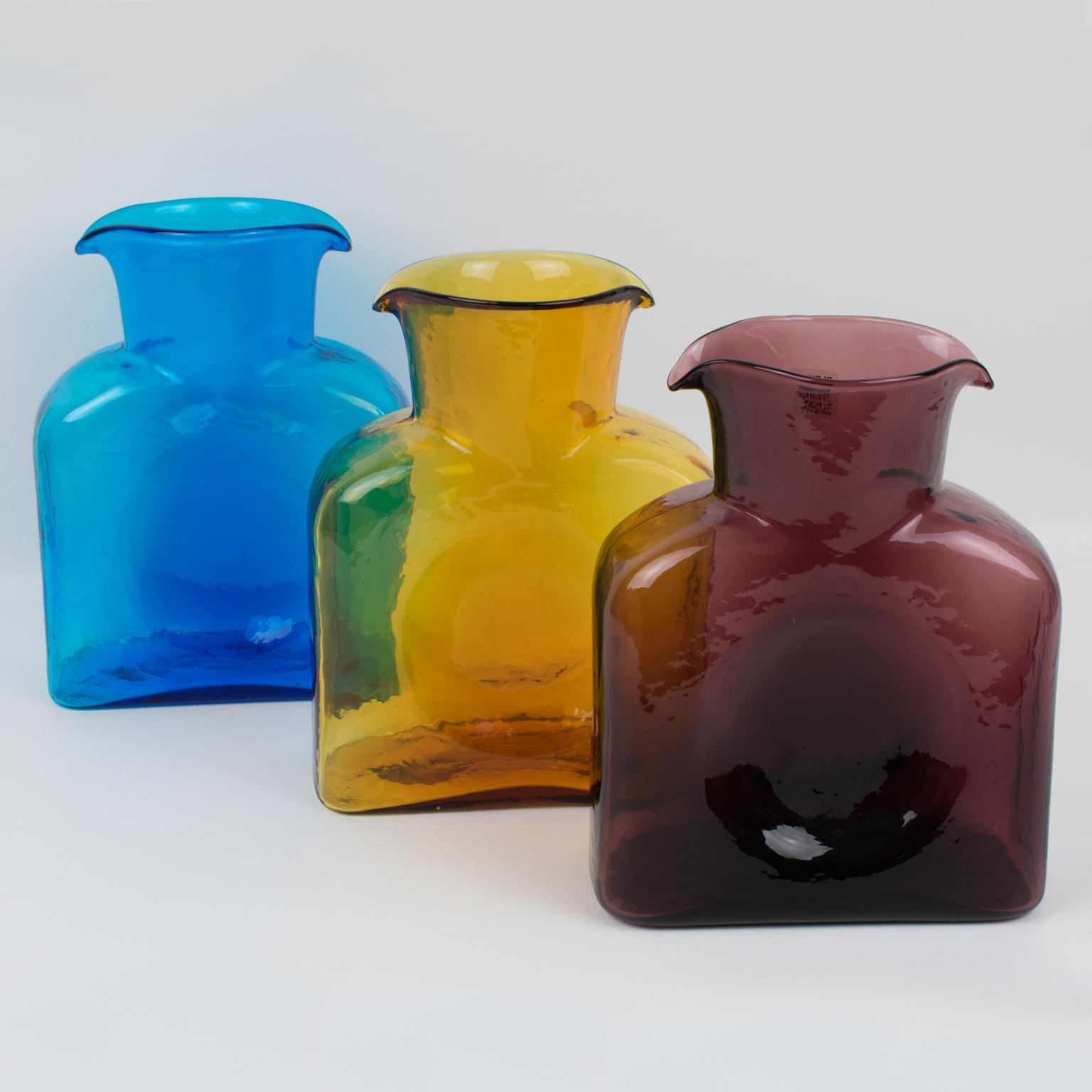 Lovely set of three pitchers, jugs, decanters by Blenko Glass Manufacturer. Square geometric shape with double spouted design. Assorted Mid-Century colors of amethysts purple, amber orange, and sky blue. The blue and the purple pitchers still have