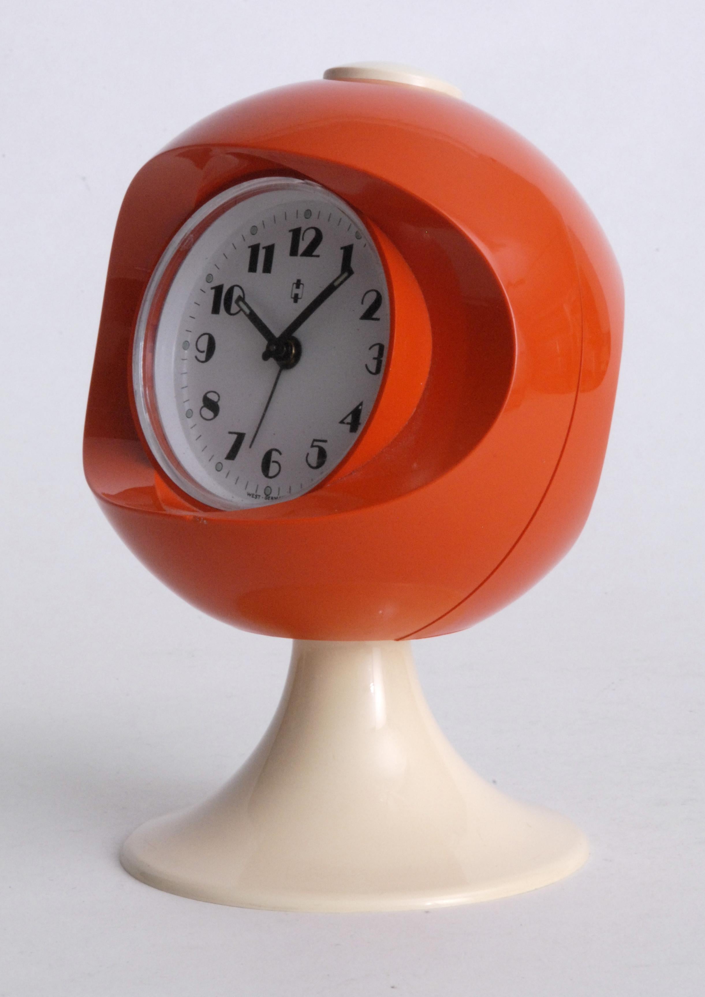 A space race influenced clock design in the form of a helmet in orange and white. An alarm clock with separate spring mechanisms for the time and alarm winding.