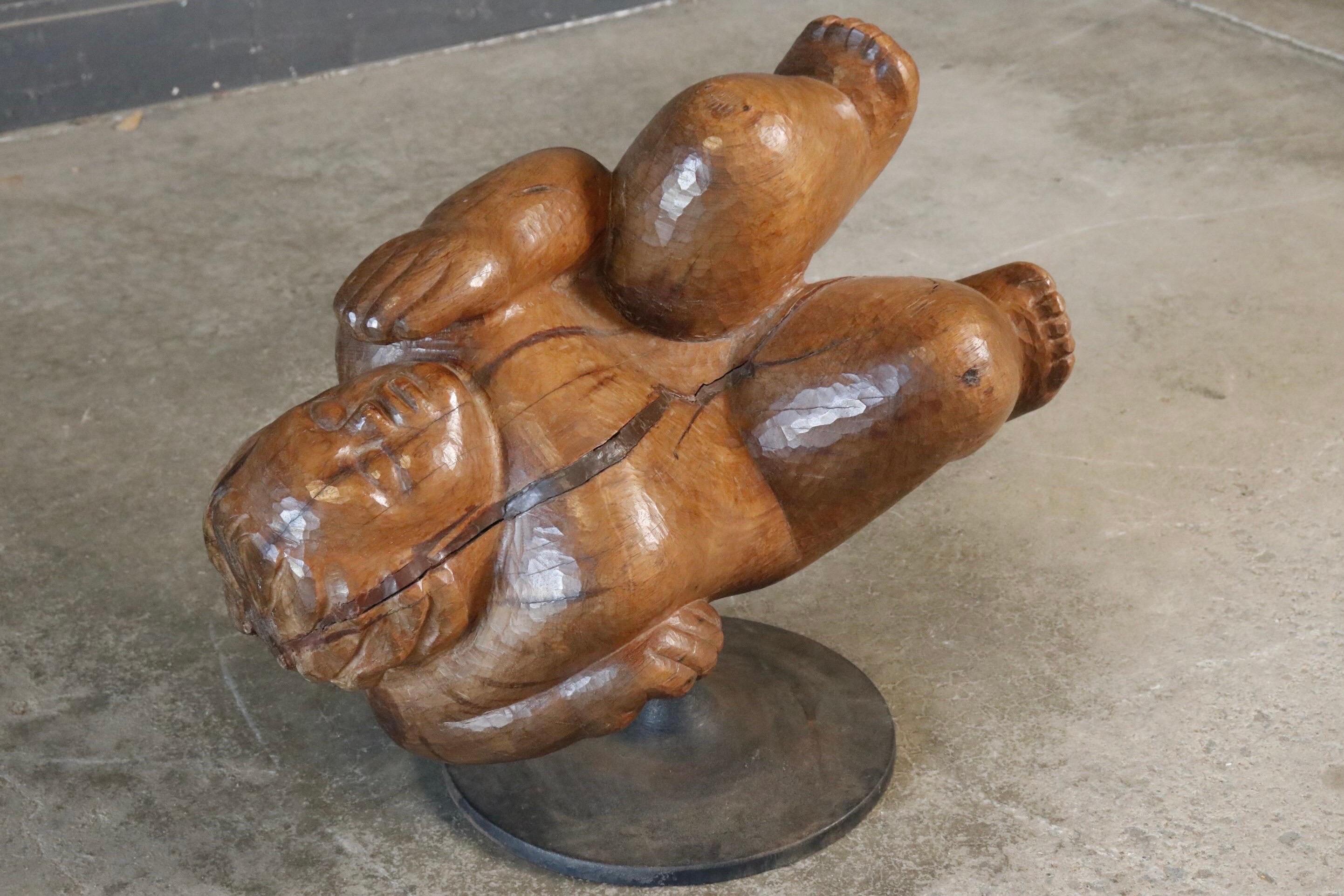 Un personnage en bois sculpté à la main est allongé comme s'il dormait béatement. Lourde et fabriquée en teck, la pièce repose sur un socle rond en fer.