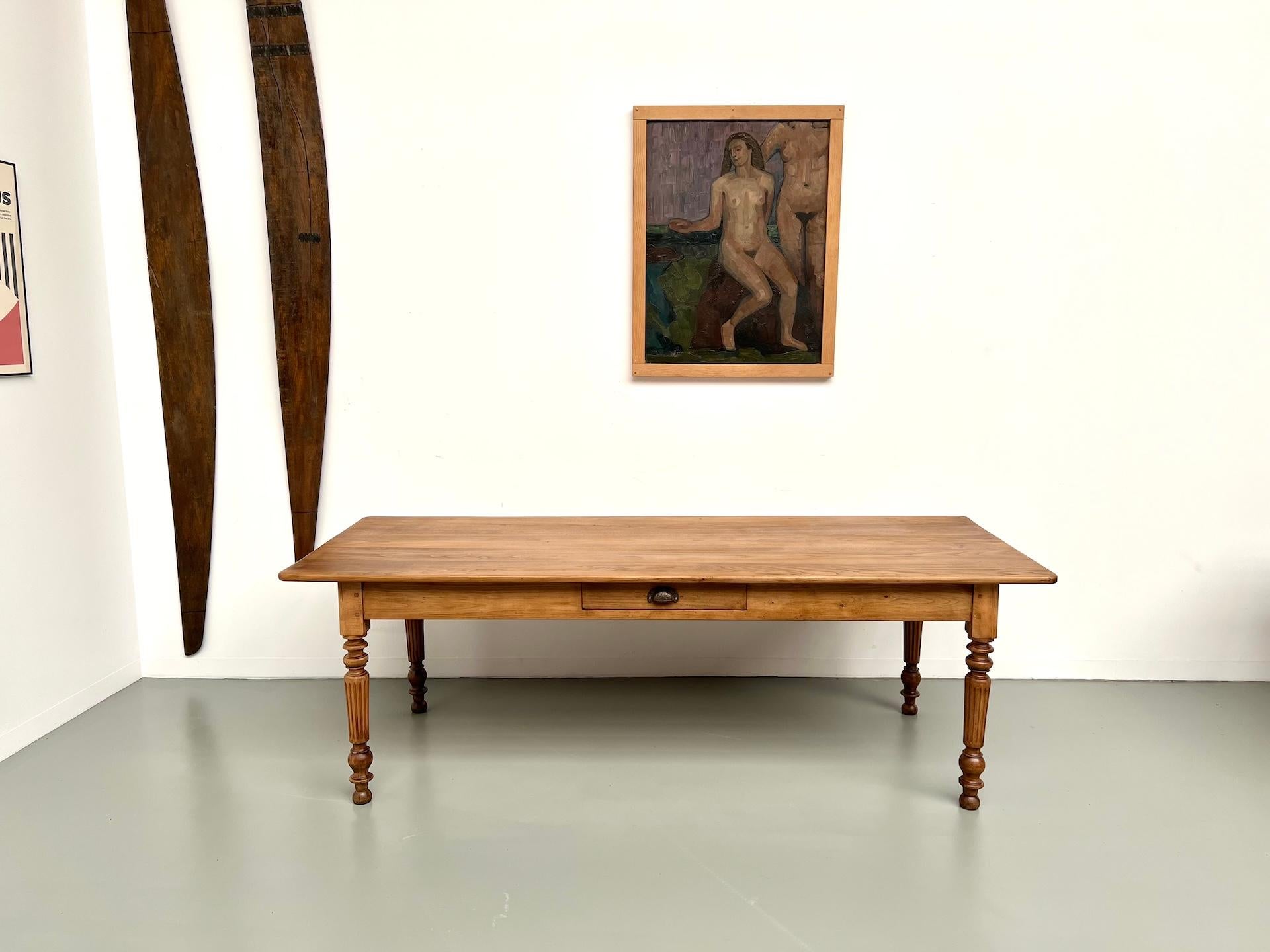 A soft, sun-washed wood, with warm golden tones and a naturally flowing grain.
The elongated top rests on four precisely turned, fluted legs – refined yet grounded.

A discreet drawer with its original shell pull sits at the center, ready to hold