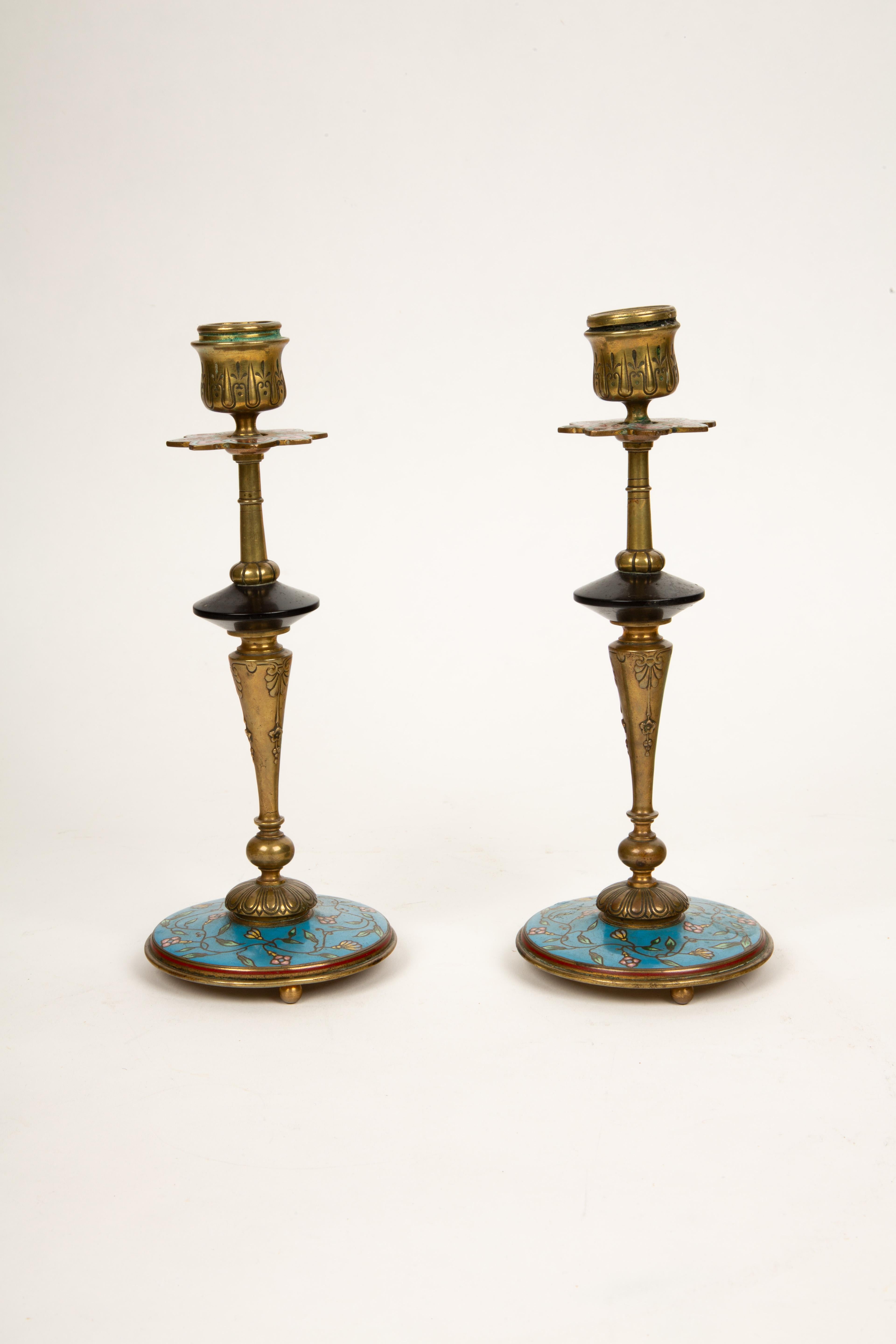 This striking pair of 19th century antique bronze candlesticks features vibrant polychrome cloisonné enamel with intricate floral and foliage motifs. Resting on stepped circular bases with raised feet, the bases are richly decorated with a vivid