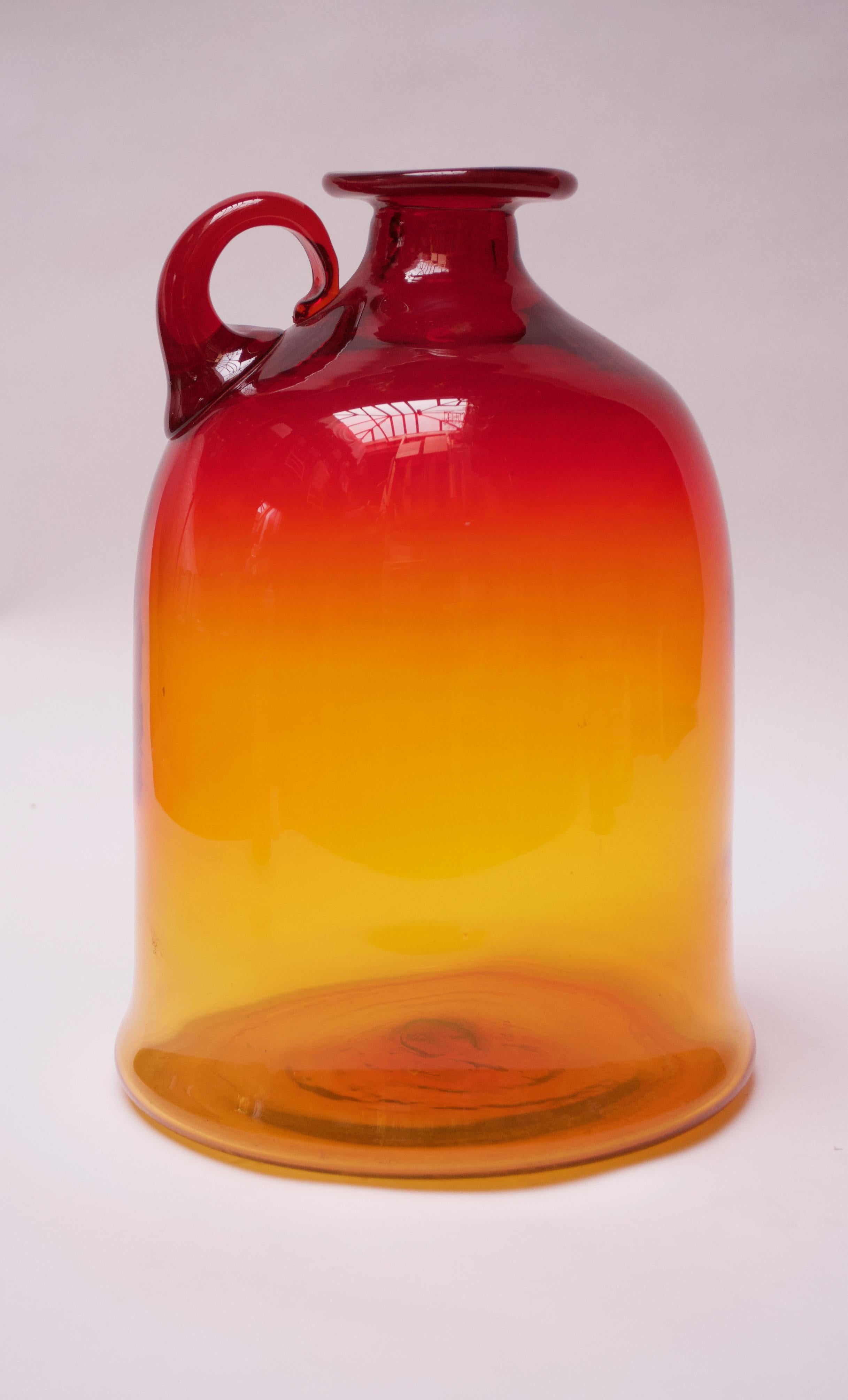 Blown glass jug designed by John Nickerson and manufactured by Blenko. This design first appeared in the 1972 Blenko catalog as model number 7226S. Large size and attractive amberina / tangerine palette with red and yellow meeting to produce an