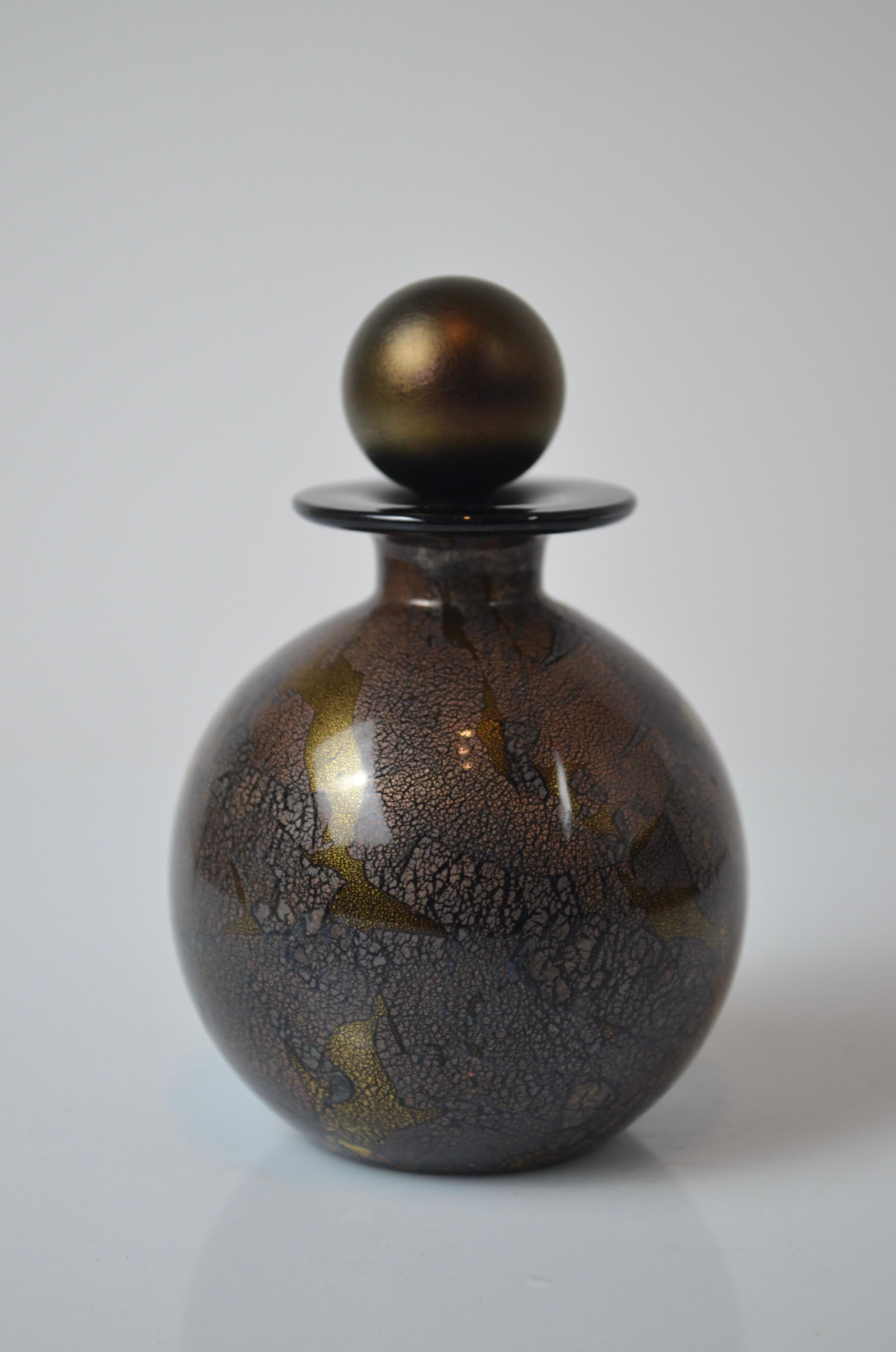 Llamativo frasco de perfume de vidrio soplado, obra del conocido Studio A de la Isle of Wight, fundado por Michael Harris.
Esta pieza presenta una rica paleta de colores oscuros -negro, cobre y dorado- con una espectacular superficie craquelada y