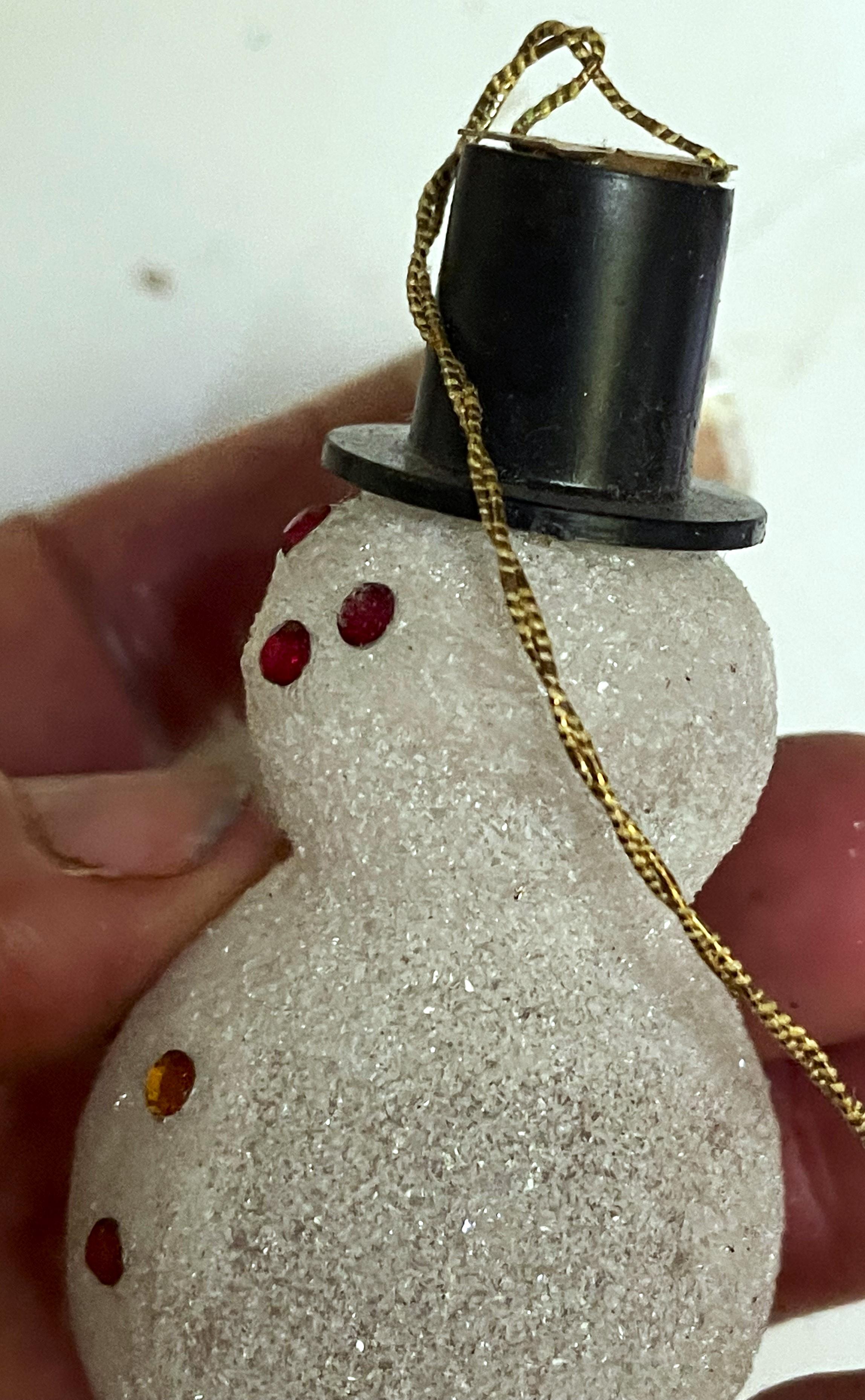 This is just the cutest little guy made of blown glass covered in a sugary snow like finish and wearing a plastic top hat with a star on top. He has rhinestones glued to his face for eyes and a mouth and as buttons down his front but one of the eyes