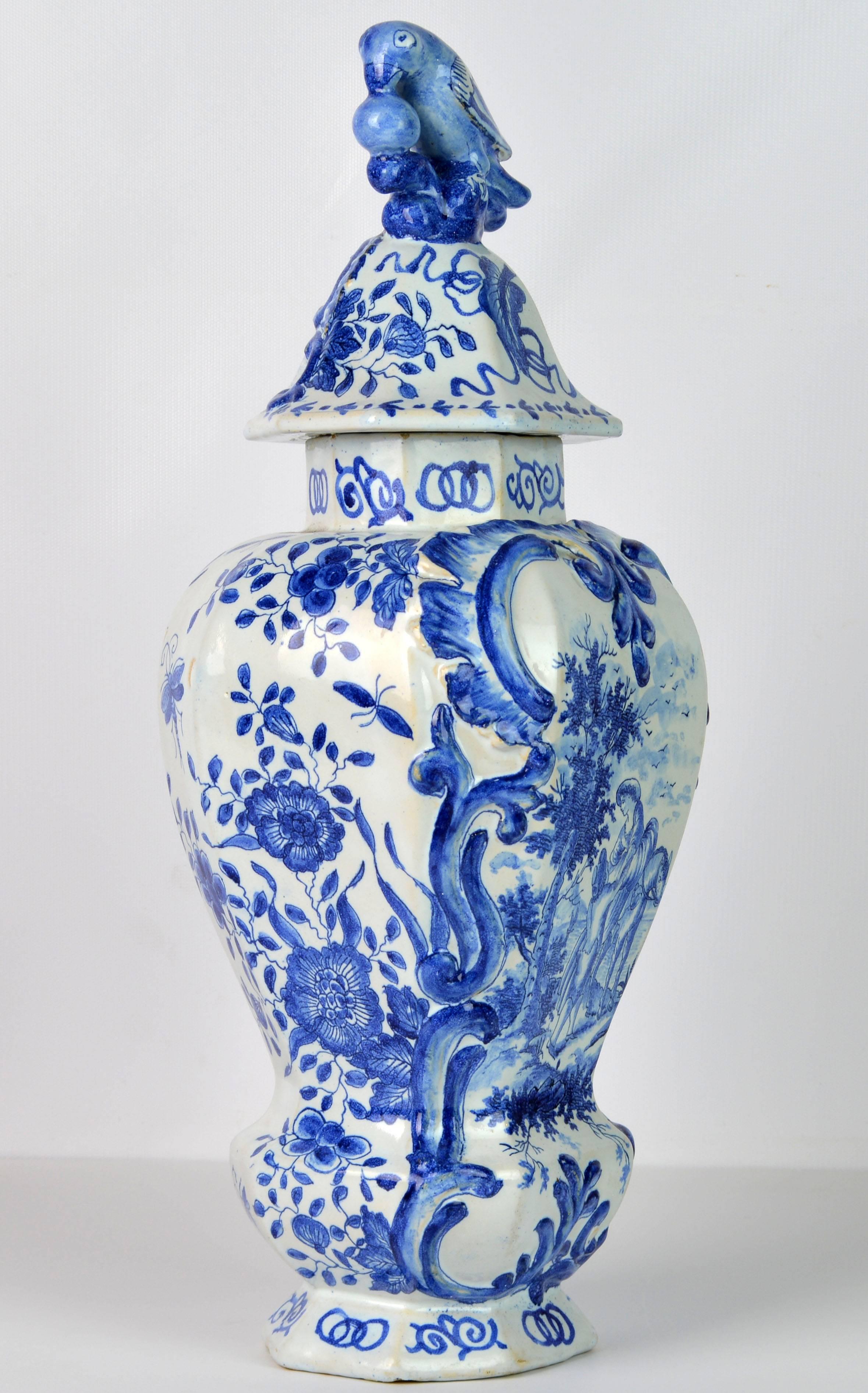 Standing 18 inches tall this lovely delft pottery jar features an octagonal cover with a bird and a berry on top above the main baluster shape octagonal body decorated with a landscape cartouche with people and a horse surrounded by flowers and leaf