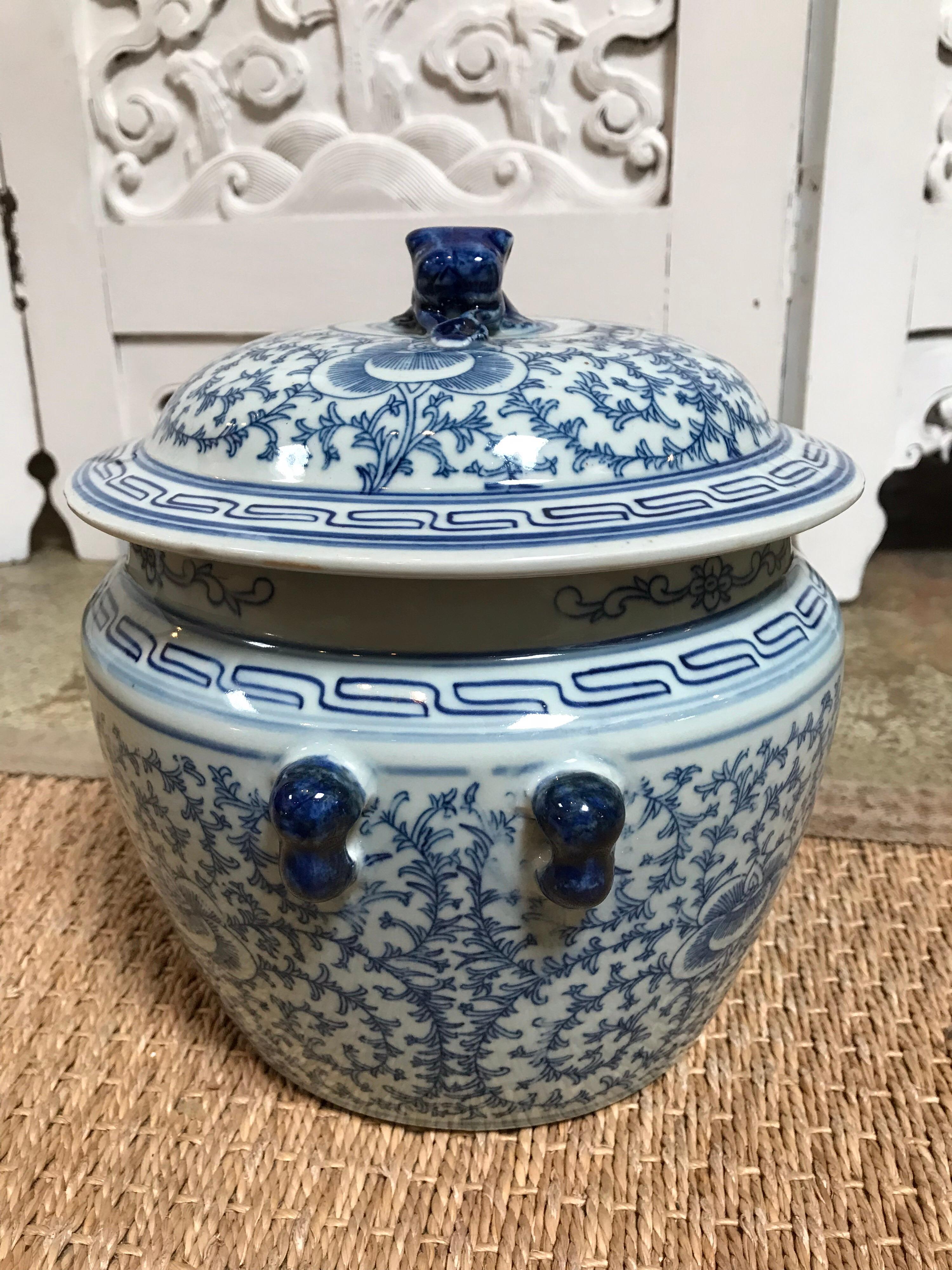 This blue and white ceramic Chinese pot is decorated in Classic blue spiraling vines sprouting from a center flower on a white background. There is also an animal figure laying down atop the lid with blue round handles on either side of the pot.