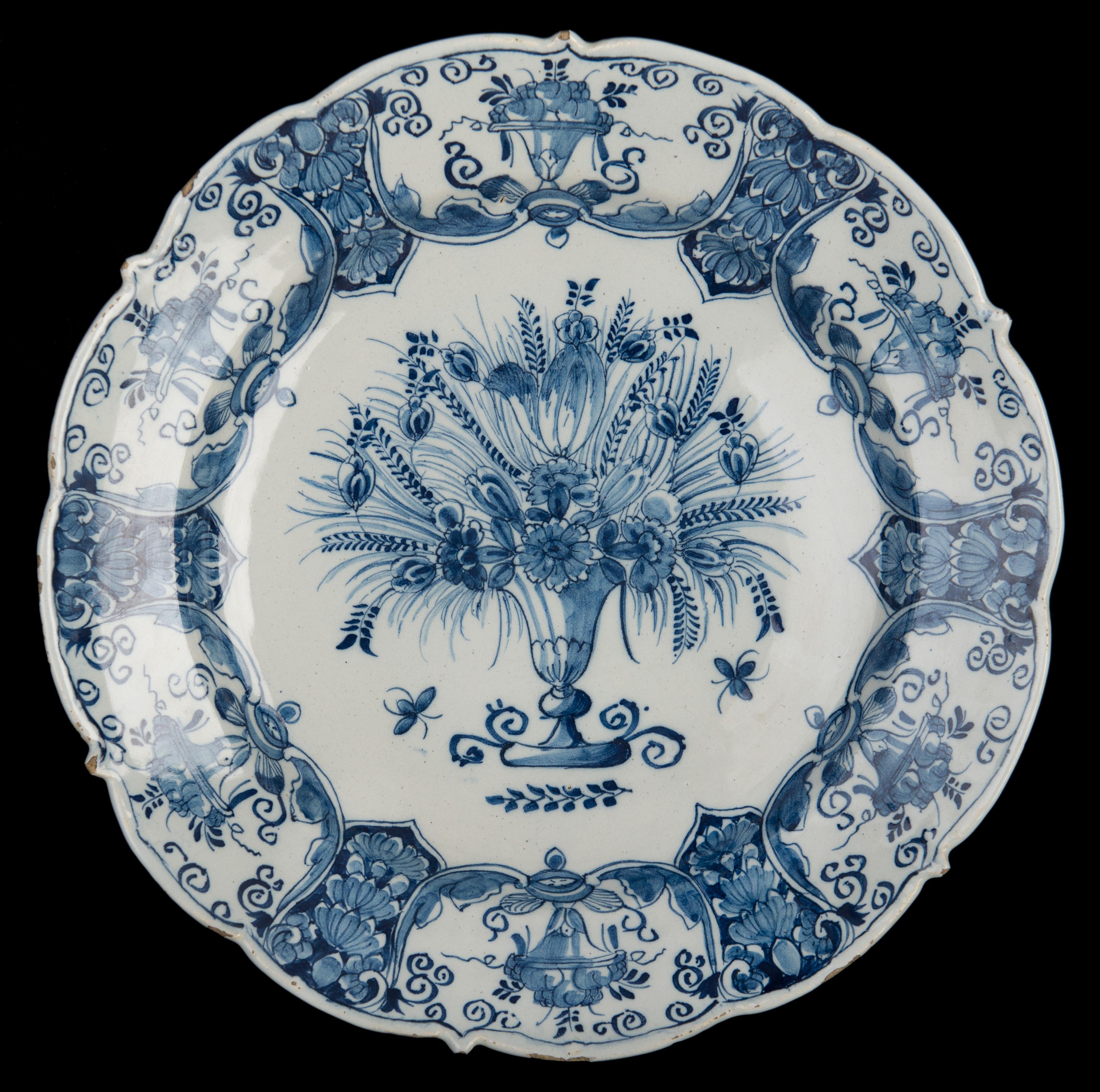 Blue and white dish with flower vase Delft, 1740-1770
The Three Bells pottery
Mark: three bells

Dish with a scalloped rim painted in blue showing a vase with several flowers and branches. A small branch is depicted under the vase. The rim is