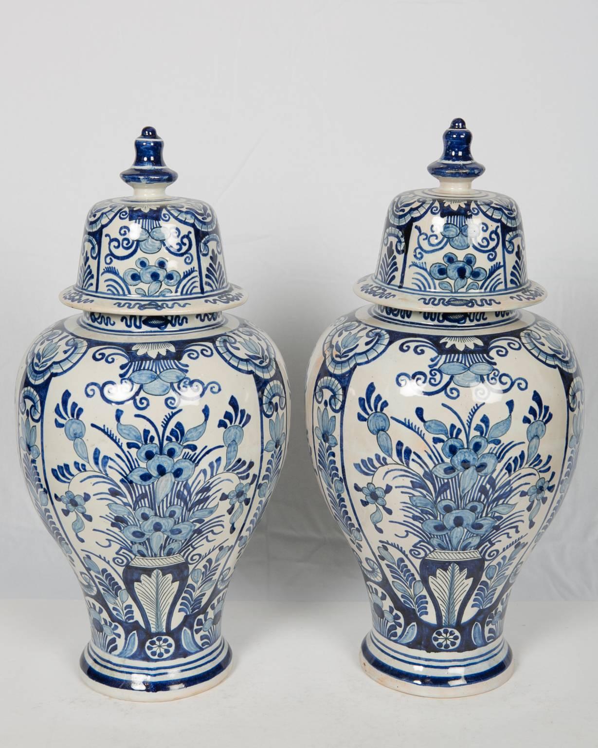 A pair of Dutch delft blue and white ginger jars decorated with panels showing a vase overflowing with flowers. The variety of contrasting blues from light to dark cobalt is what makes this elegant pair of vases so wonderful. The covers are