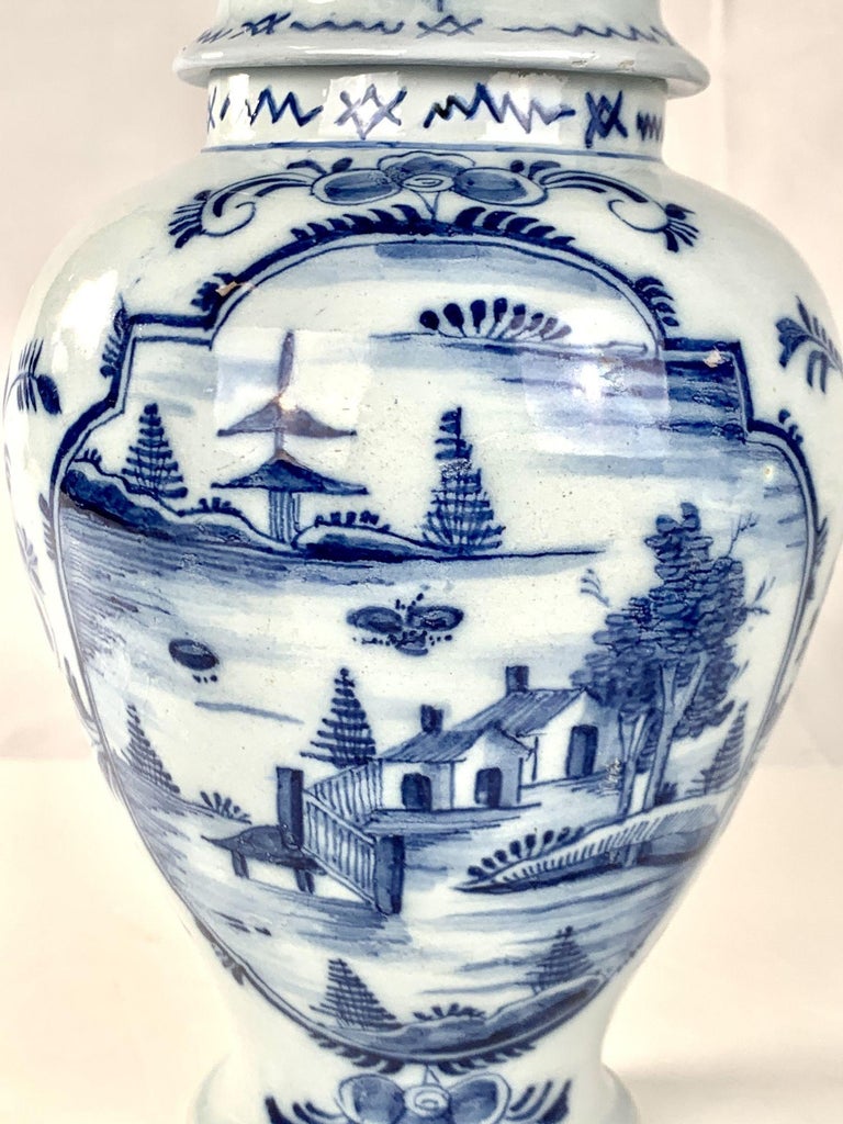 This charming blue and white Dutch Delft jar is decorated in shades of cobalt blue.
Hand-painted in the late 18th century circa 1780, the jar mixes rococo and chinoiserie design elements.
We see a modest house with blossoming fruit trees and pine