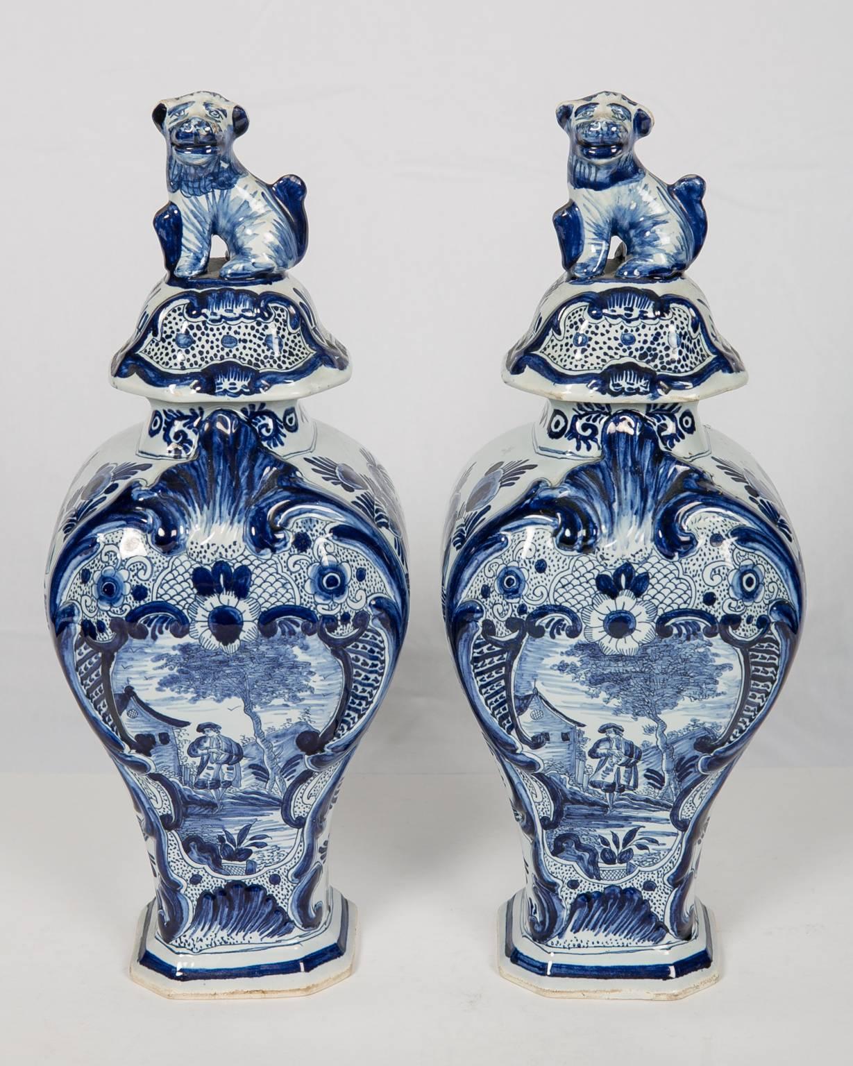 A pair of Dutch Delft blue and white mantel vases painted in particularly deep cobalt showing a nobleman walking on a country road. To his left we see a Dutch farmhouse, to his right a leafy tree. The scene is framed by a symmetrical design of