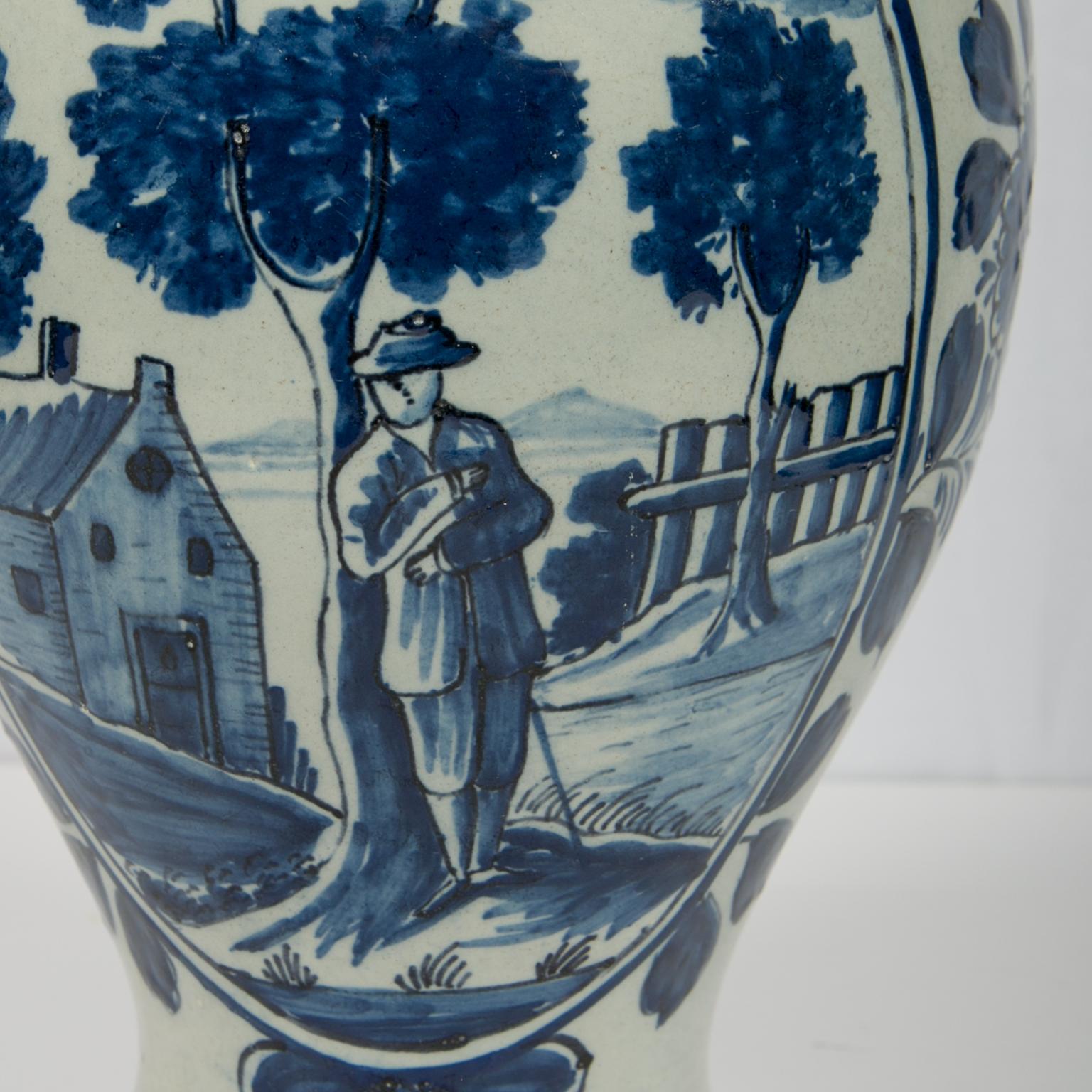 A blue and white Dutch delft jar painted with a simple country scene featuring a gentleman holding a walking stick and resting against a tree. The tree and the trees nearby have sponged decoration defining their leaves. The clouds in the sky are