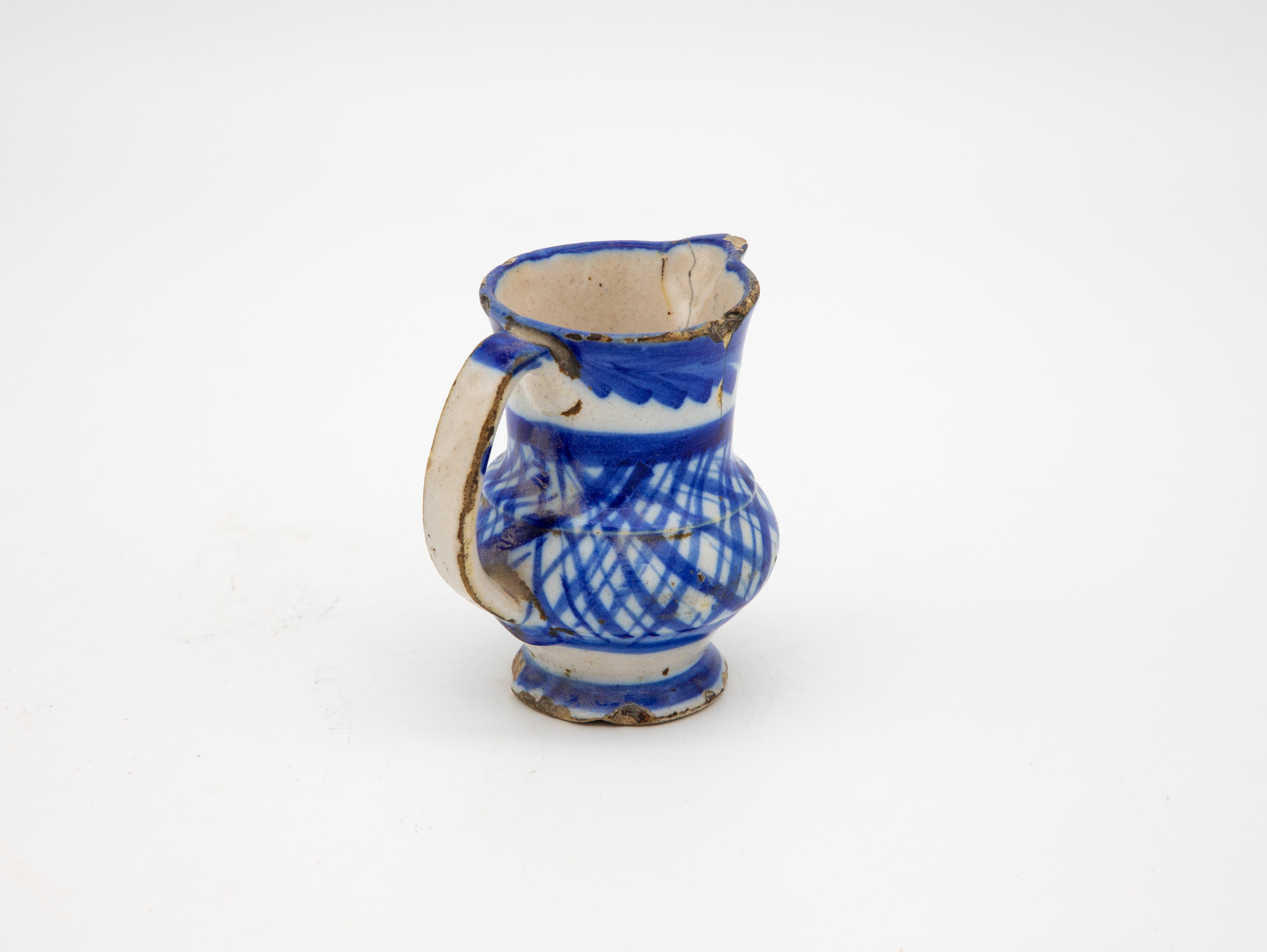 Un petit pichet ou une cruche en céramique blanche avec une décoration à rayures bleues et blanches et des hashmarks. Petits éclats de perte à la base et au sommet. Ce petit pichet est d'origine inconnue, mais probablement espagnole. Fin du 19e