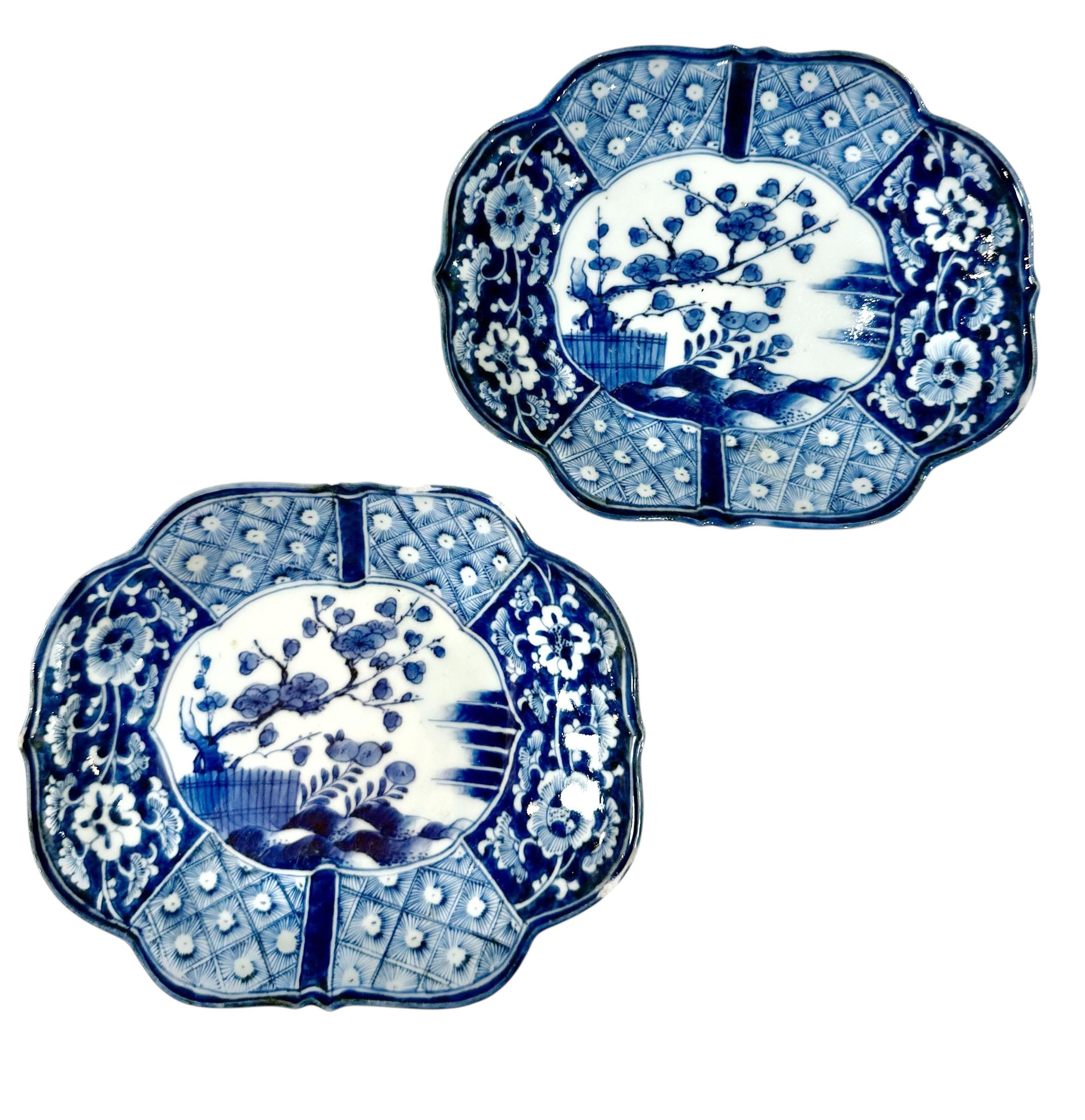 Un charmant ensemble de quatre plats en porcelaine japonaise, magnifiquement peints à la main en bleu et blanc traditionnels. Le dessin central représente un paysage clôturé avec un arbre et des cerisiers en fleurs, rendu par un délicat travail de