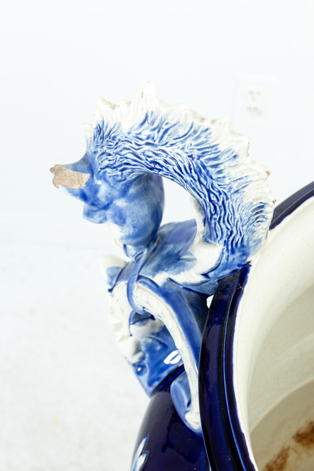 Blue and white painted Majolica urn on stand detailed with neoclassical motifs such as acroterion on the base, scrolled arms on the urn, and gadroon trim. Please note of wear consistent with age including minor finish loss and chips.