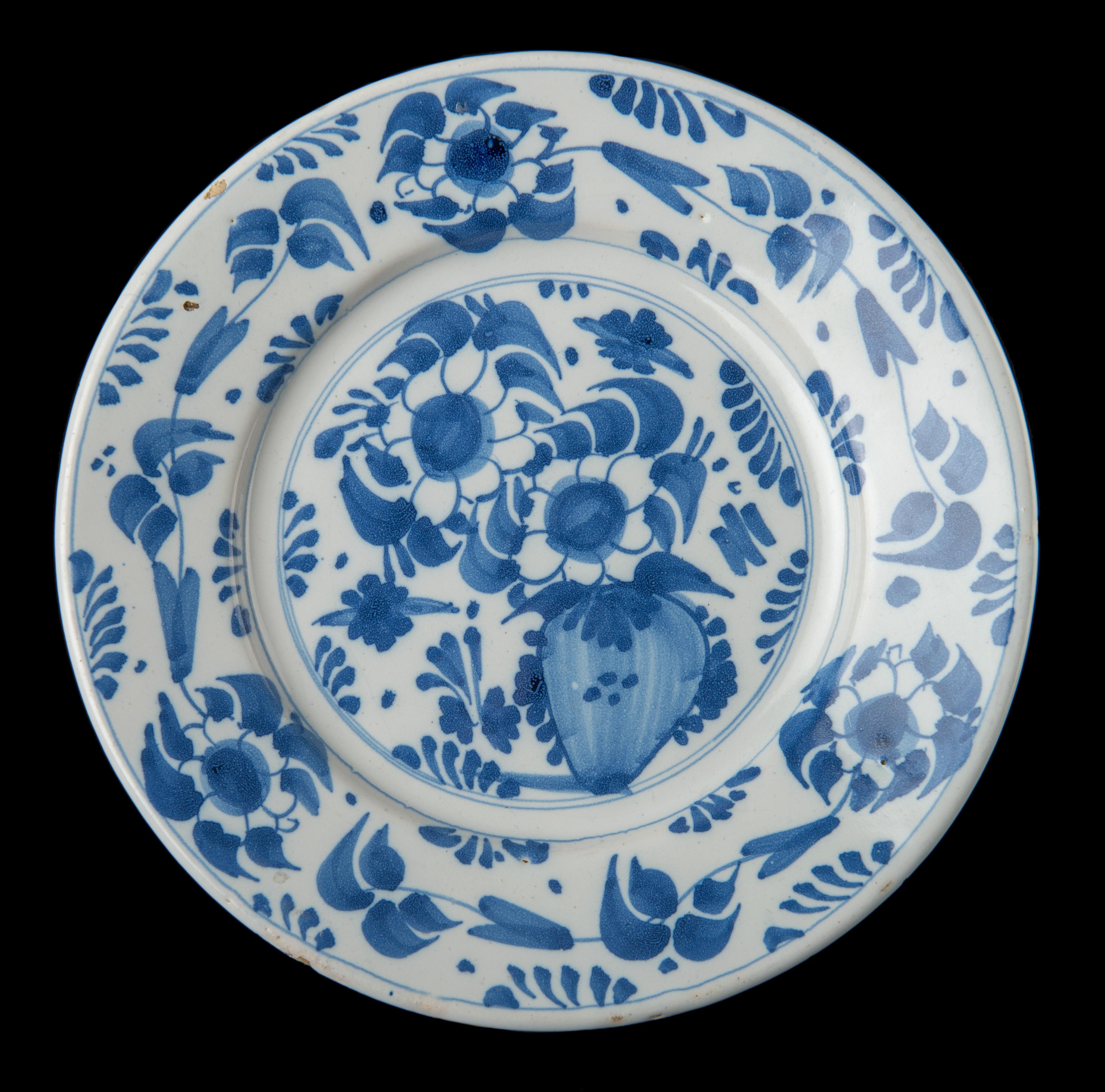 Blue and white plate decorated with of a vase with two flowers in a double circle. The well and flange are decorated with a continuous frieze of flowers, foliage and branches of leaves. The plate has no foot rim. 

The decor of a vase with two