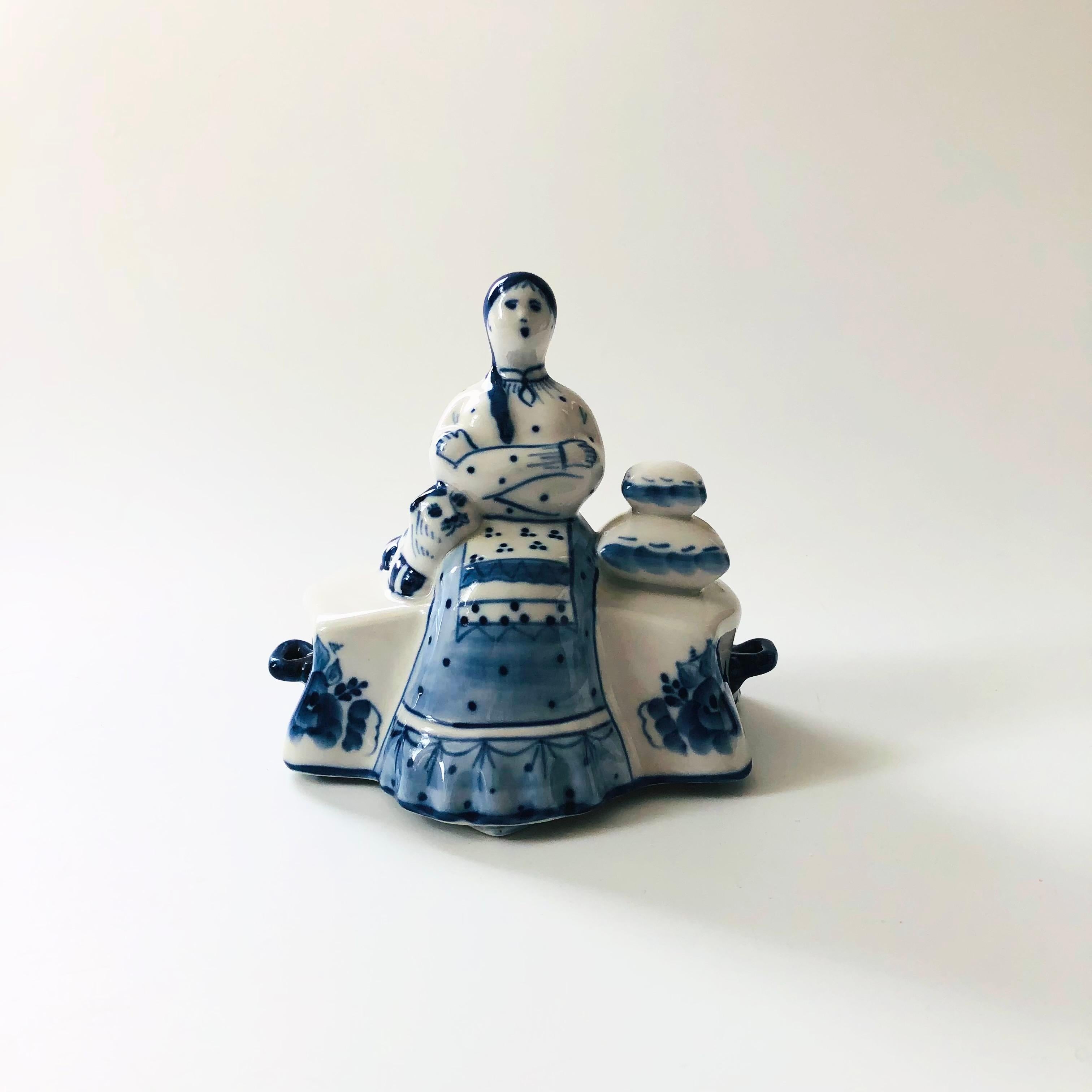 A vintage porcelain butter dish in the shape of a woman. Blue and white glazes with beautiful hand painted detailing throughout. A cat sits on the left side of the woman and a stack of pillows sits to the right. The top lifts off to reveal the dish