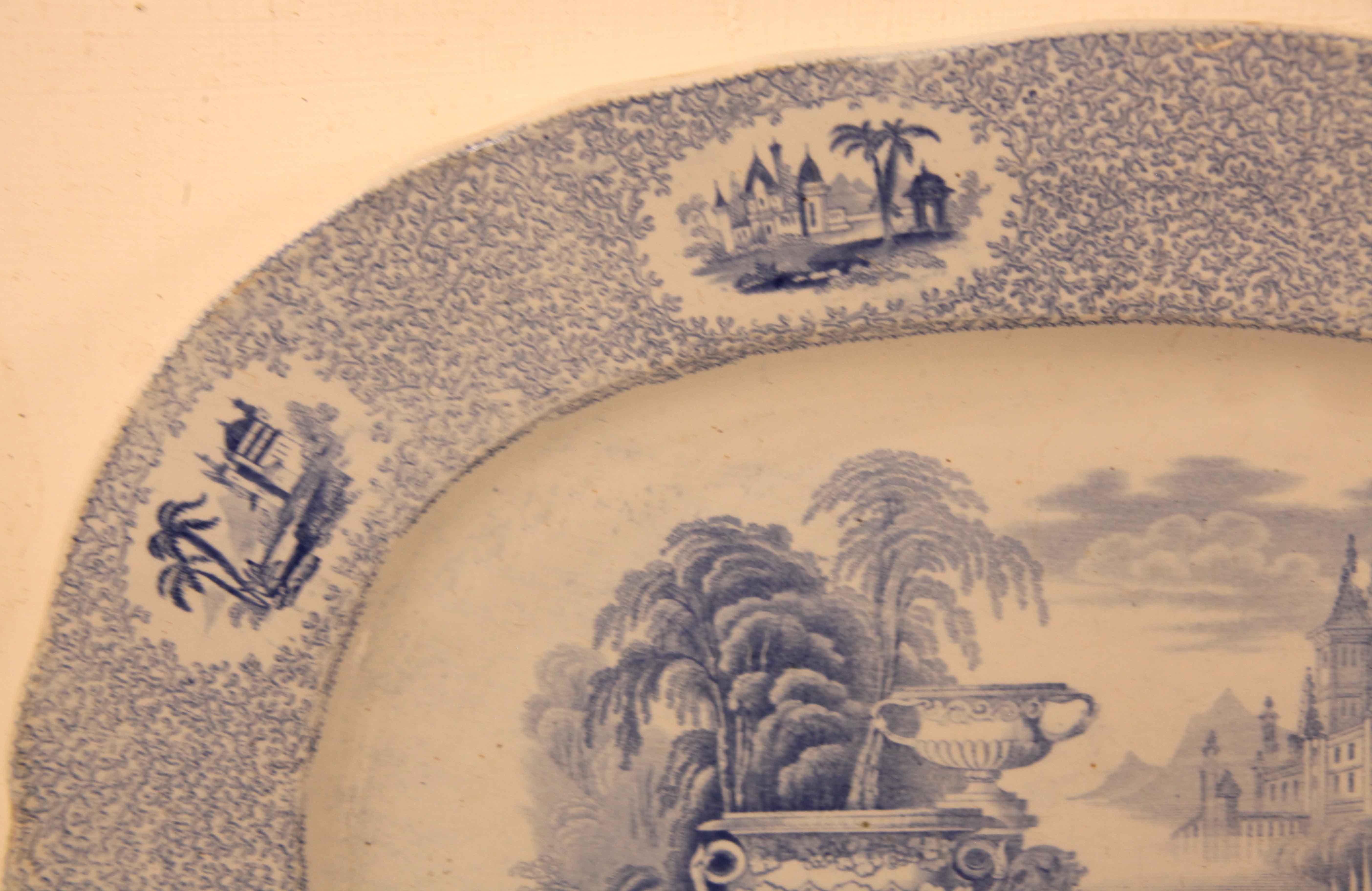 Blue and white Staffordshire platter,  the border of this platter is a dense stylized foliage interspersed with oval cartouches of various middle eastern scenes.   The main body features a garden scene with multiple footed urns, nymphs,  flowers, 