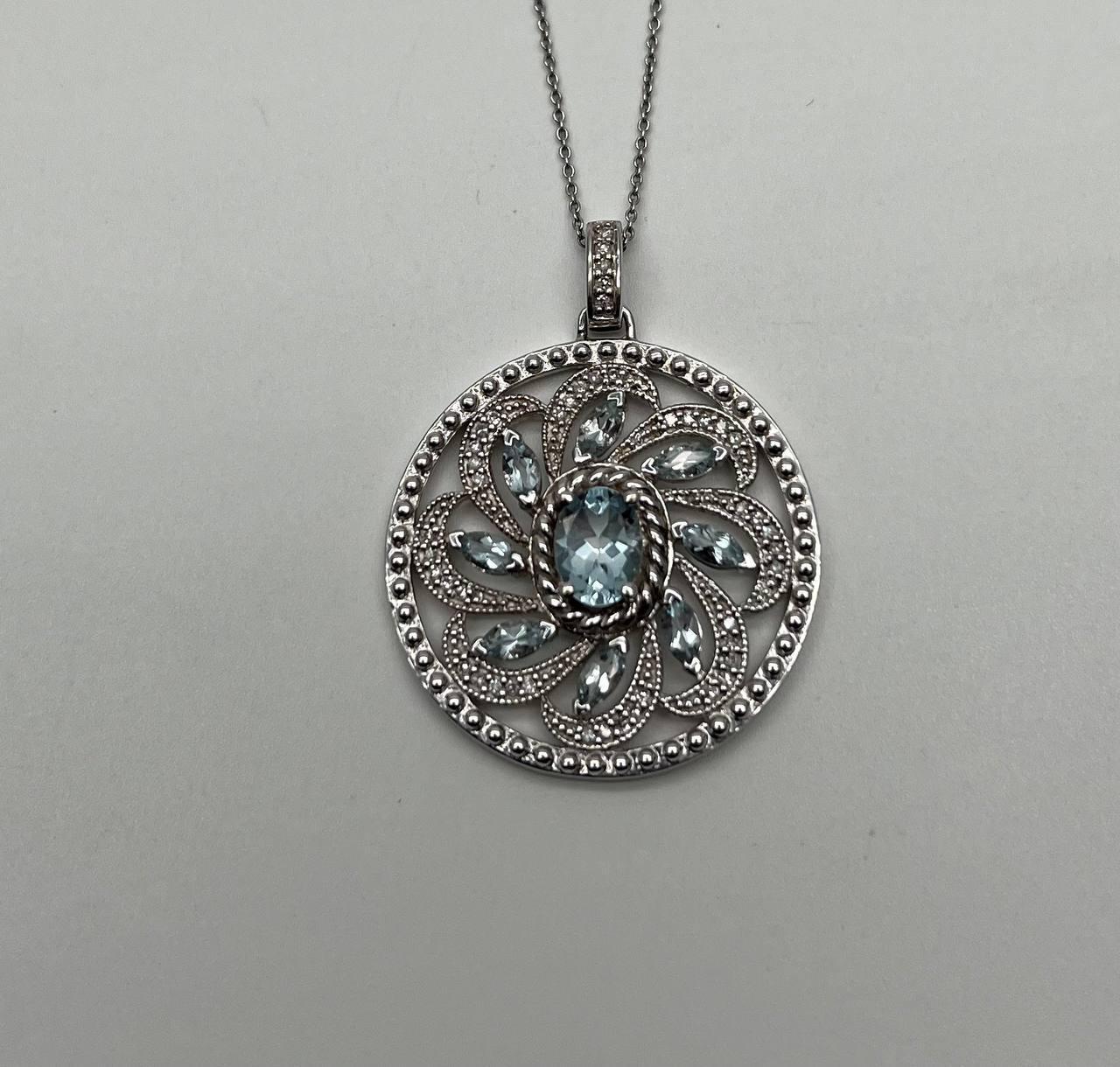 This intricate pendant necklace is crafted with precision and artistry. The design features a circular motif with a central oval blue aquamarine, surrounded by a pattern of smaller marquise-cut blue aquamarines. The stones are set within a detailed,