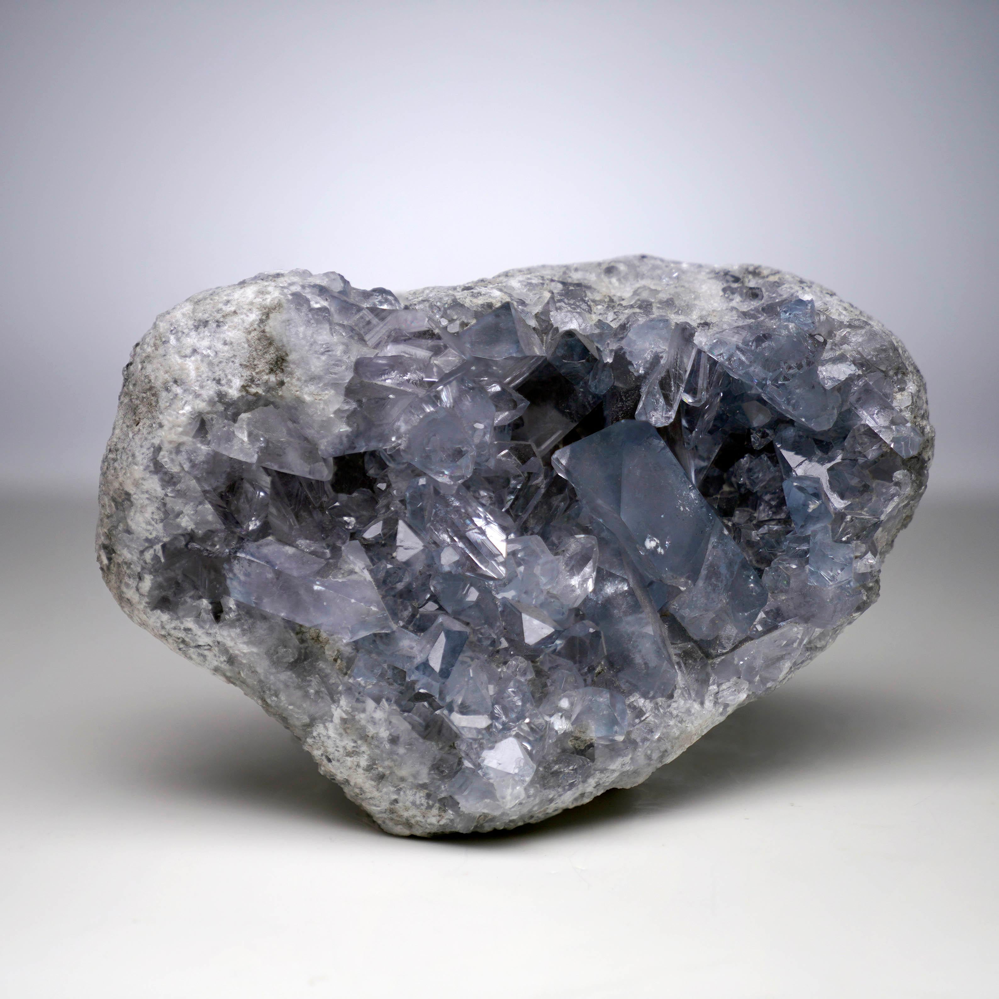This Blue Celestite geode from Madagascar showcases a cluster of large, gemmy Blue Celestite crystals with stunning light to denim-blue pigmentation. This cabinet-size specimen of the strontium sulfate mineral displays glassy clarity, with many of