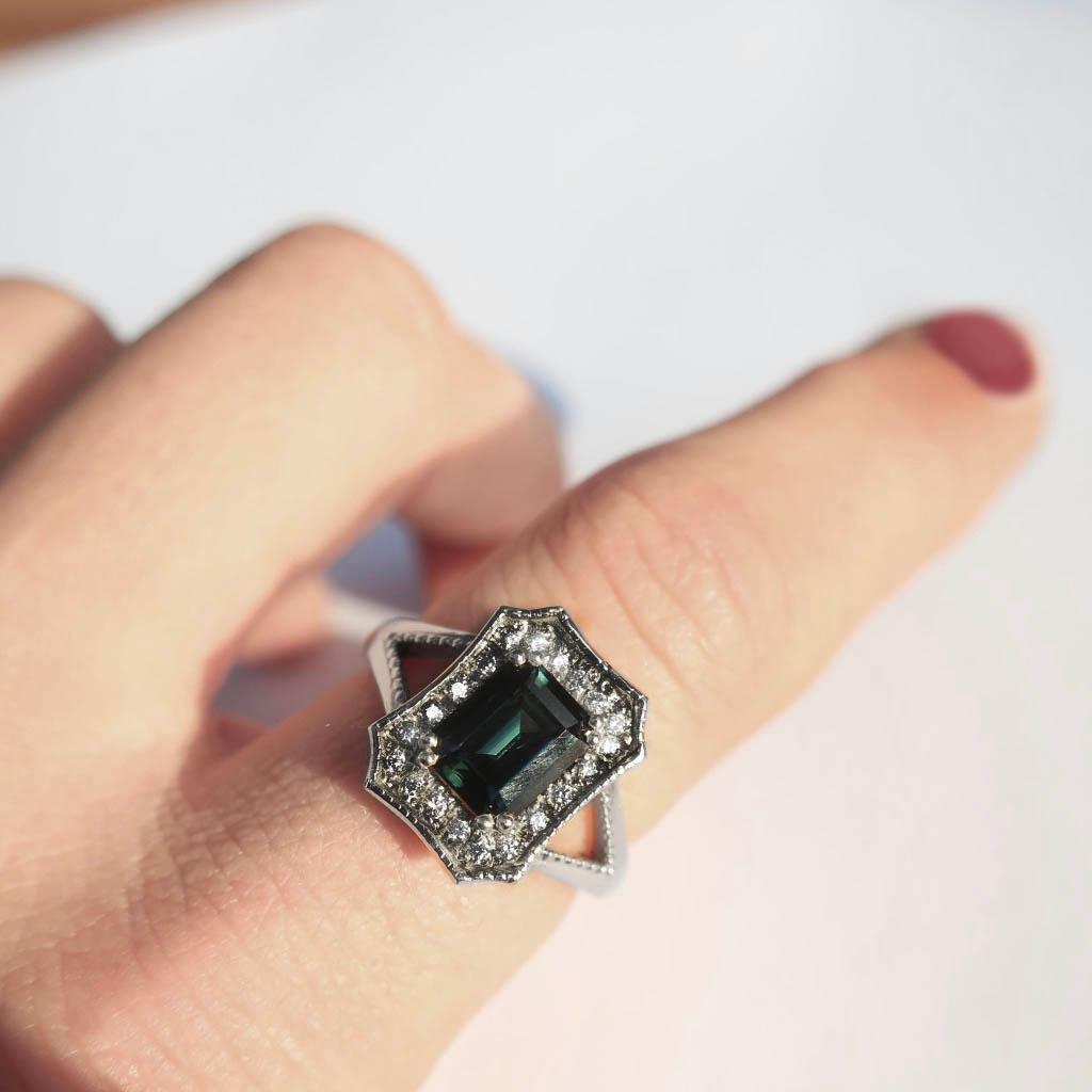 This gorgeous deep blue emerald cut ring has a halo of white diamonds on a split band. This combines classic vintage silhouettes with a modern flare making this a timeless heirloom piece. 
This deep blue/ teal sapphire weighs 1.36 carats and