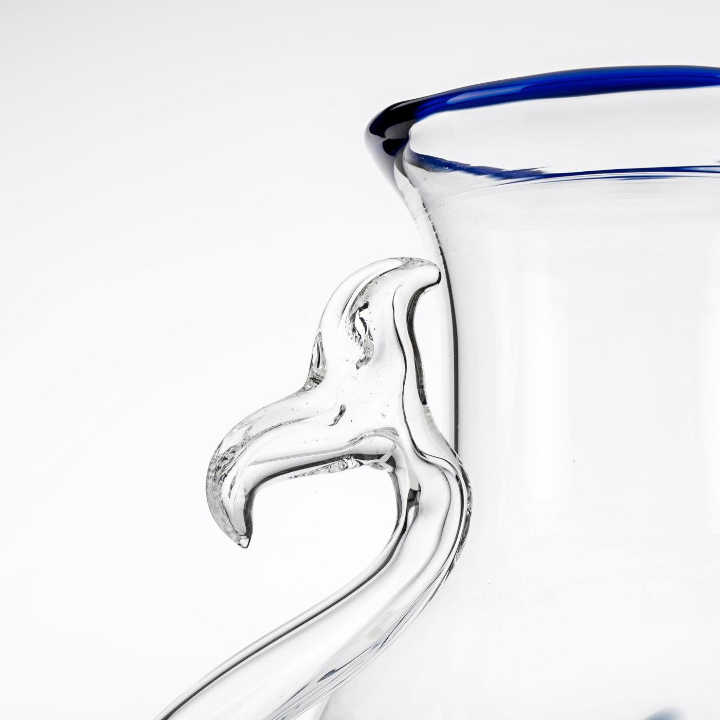 This elegant jug features a blue stylized fish scale pattern on the bottom section of its sinuously curved body, while a delicate blue glass line highlights its lip. The unique handle of the piece is shaped like the tail of a fish and curves upwards