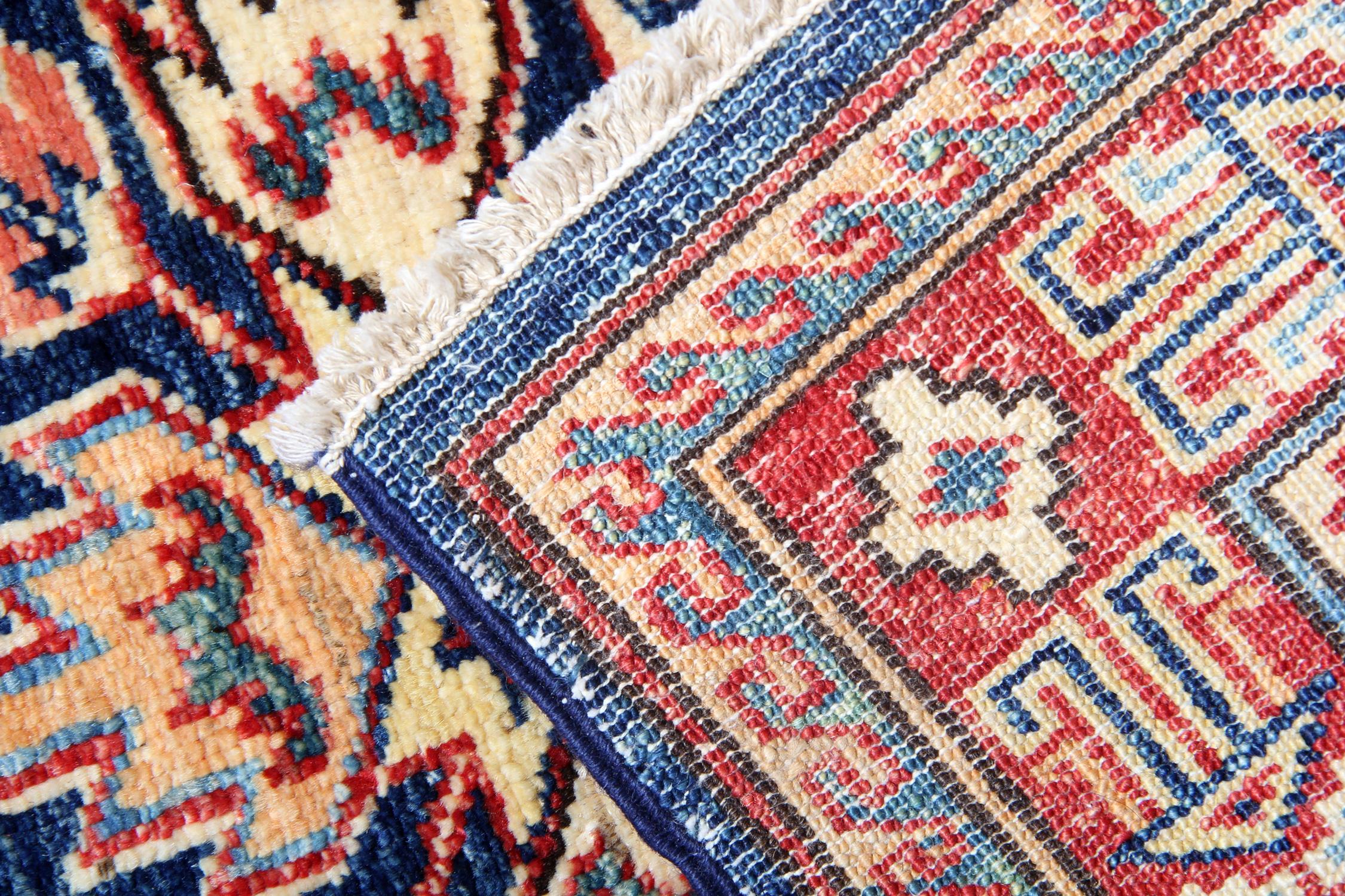A beautiful new traditional Afghan Kazak rug features three conventional tribal medallions woven in accents of red, orange, and cream through the centre of a navy-blue field. The geometric rug design is then finished with a highly-detailed repeat