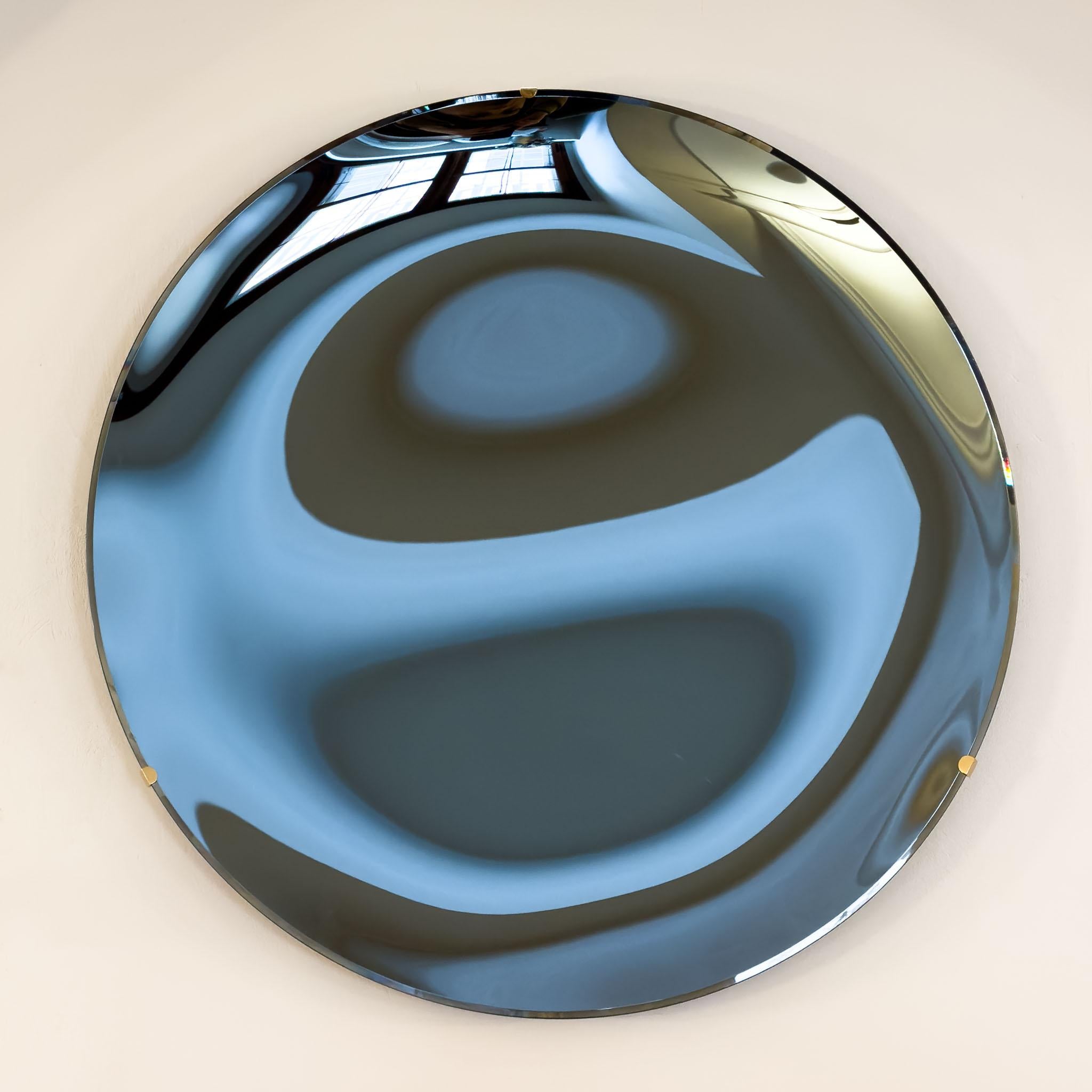 Large round wall mirror in concave shape with faceted edge. The wall mirror is made of blue glass and is held by a minimalist brass wall bracket.