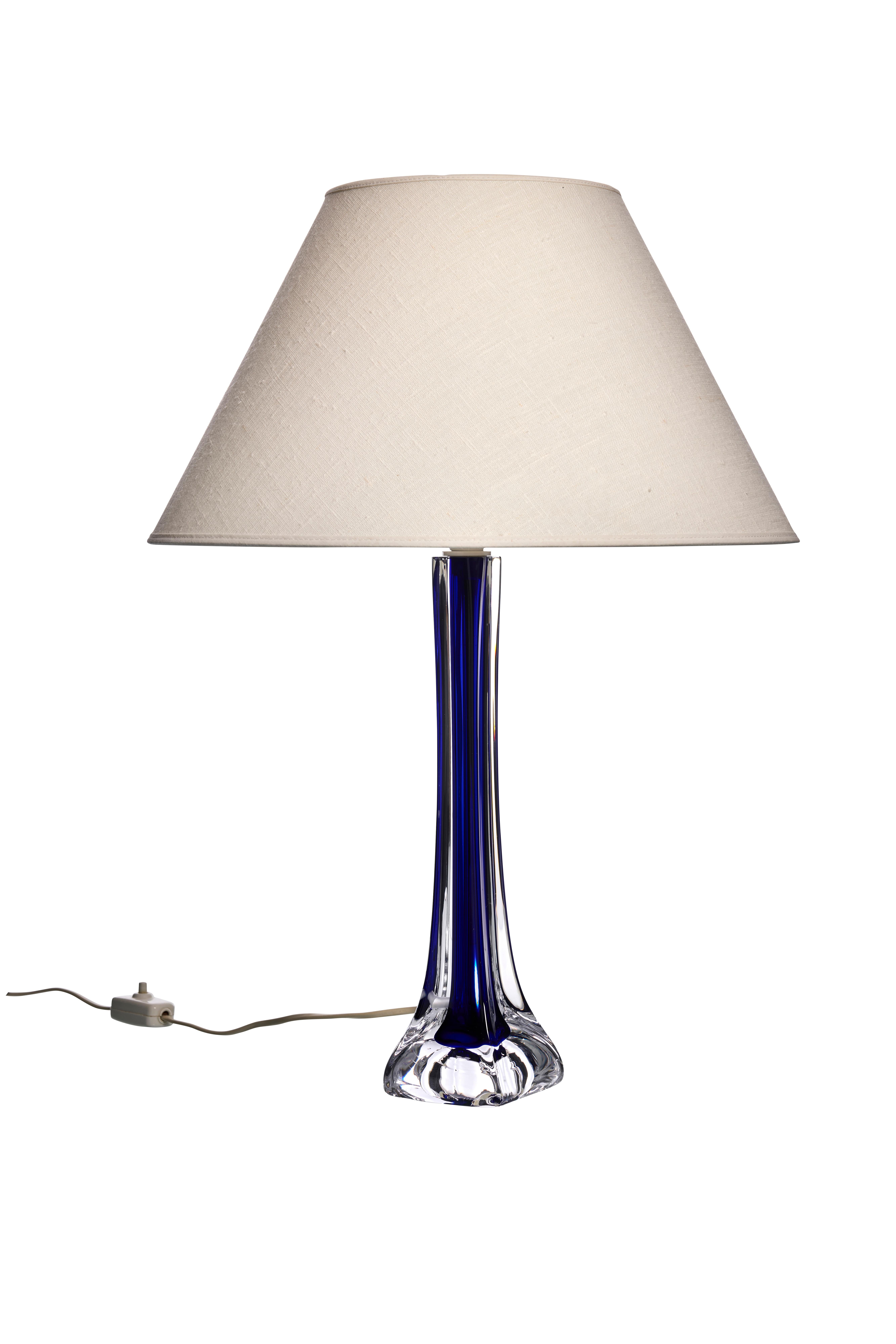 Esta elegante lámpara de sobremesa tiene una base alta y esbelta de llamativo cristal azul. El cristal transparente de la base se transforma a la perfección en un azul intenso y profundo, creando un efecto visual cautivador. La base está diseñada