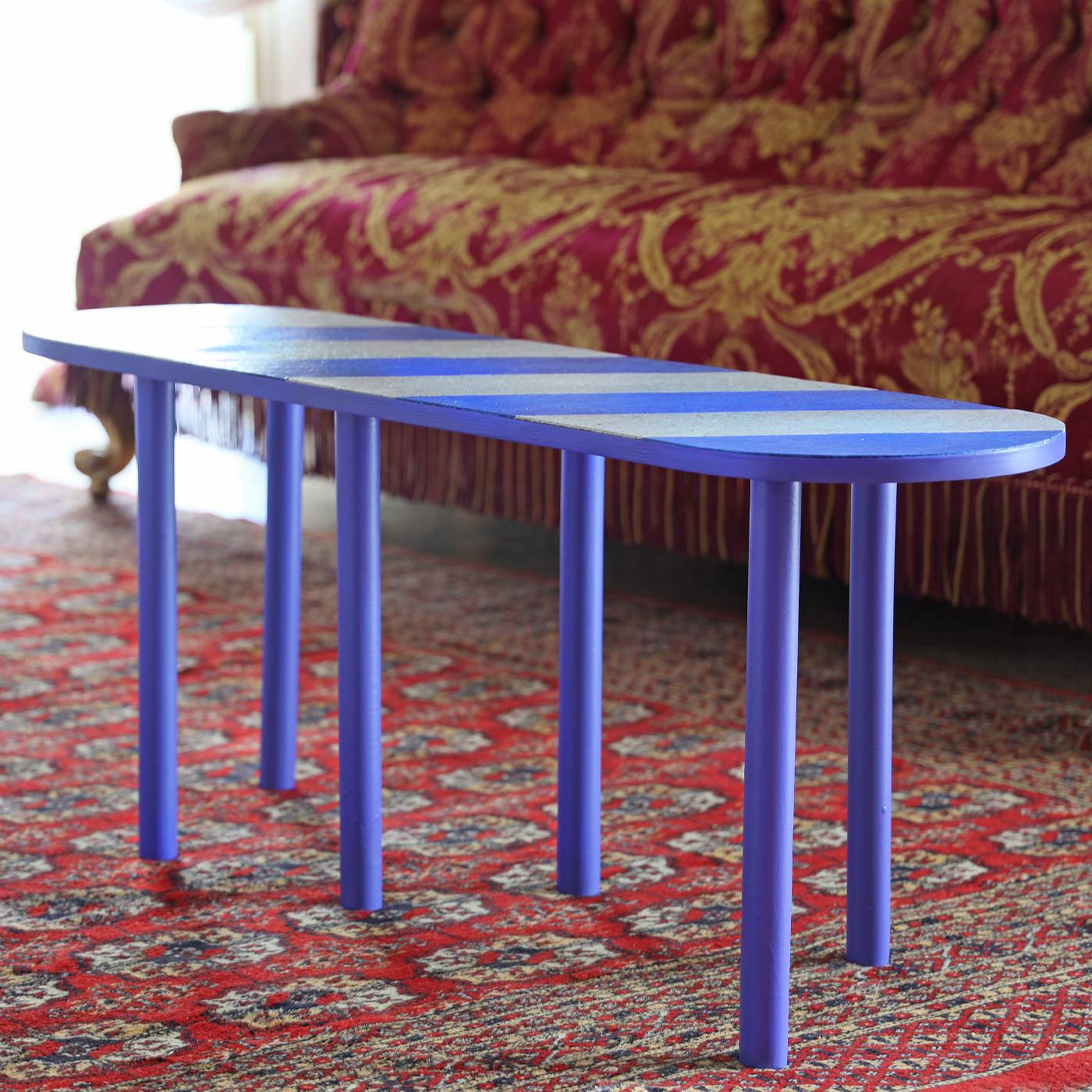 This sustainable side table strikes with its vibrant ultramarine shade painted with water-based glaze. The poplar blockboard frame comprises six cylindrical legs sustaining an elongated top with rounded extremities, whose surface displays a pattern