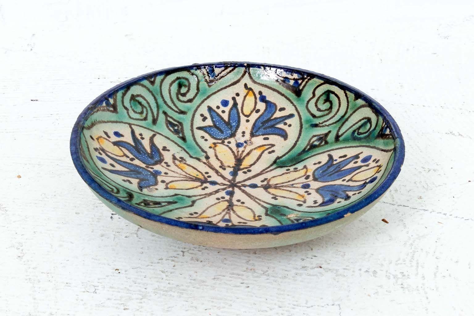 A Morrocan bowl featuring blue flowers radiating from the center. Small yellow leaves around the flowers and a light green border. Wire on back allows for wall display. 8