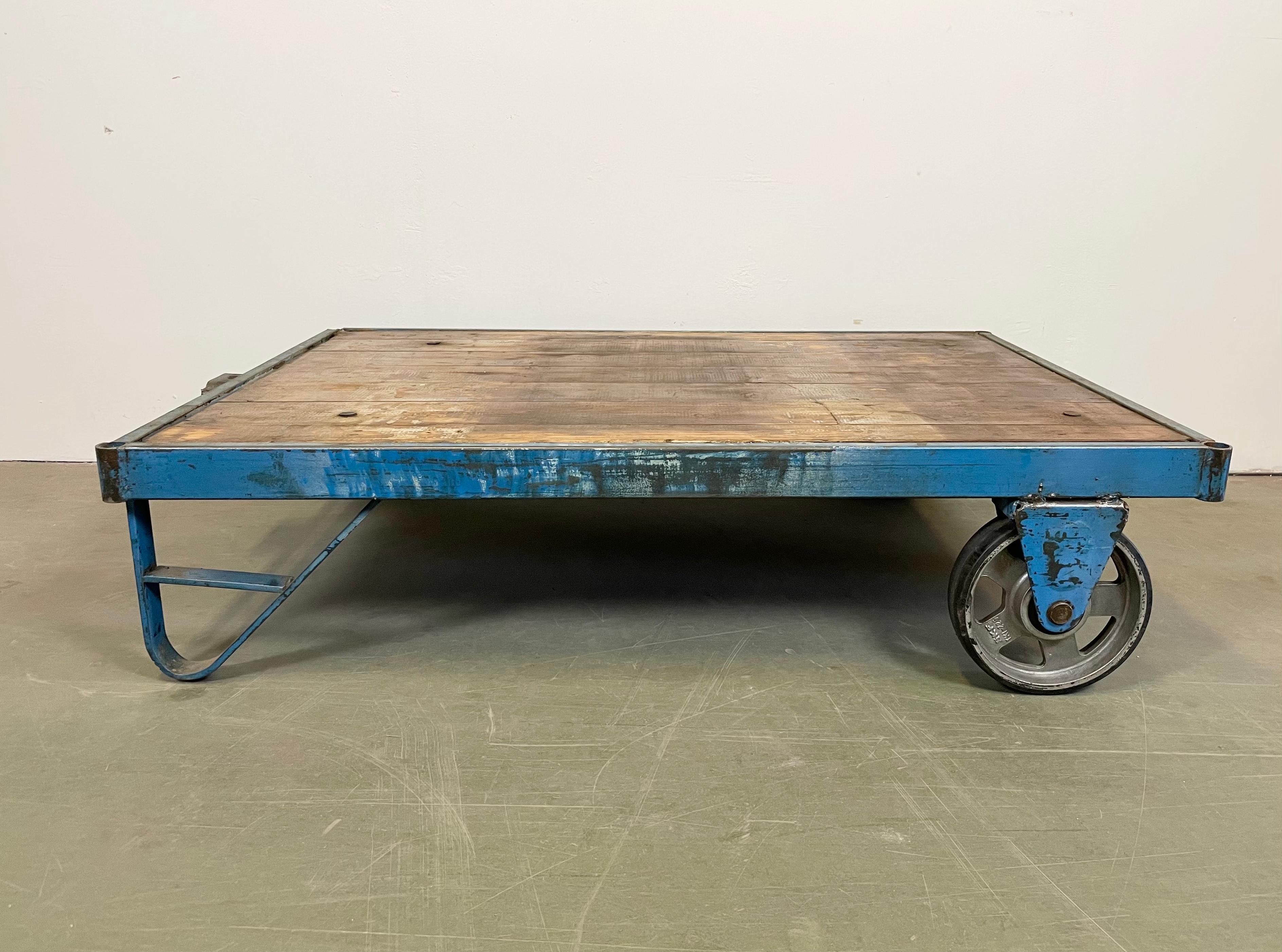 Former pallet truck from a factory now serves as a coffee table. It features a blue iron construction with two original wheels and solid wooden plate. The weight of the table is 40 kg.