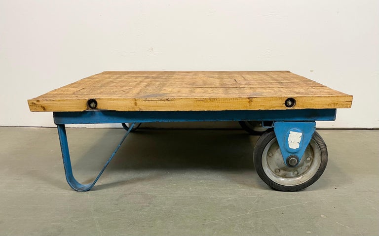 Former pallet truck from a factory now serves as a coffee table. It features a blue iron construction with two original wheels and a solid wooden plate. The weight of the table is 30 kg.