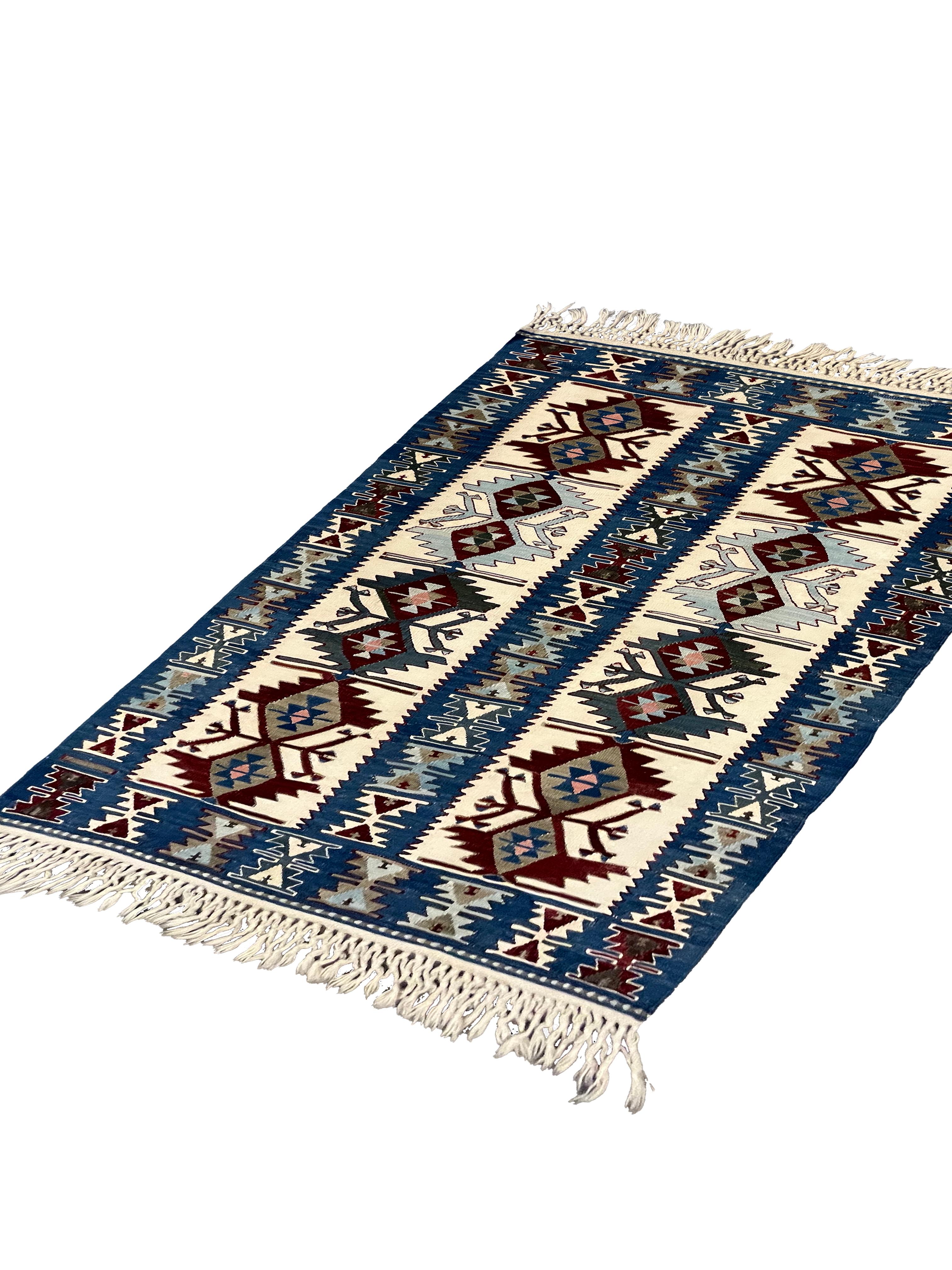 Ce petit kilim est un élégant tapis turc Kilim tissé à plat à la main. Le dessin présente un motif géométrique traditionnel, tissé symétriquement avec un grand niveau de détail. La palette de couleurs comprend des accents de bleu, de crème, de rouge