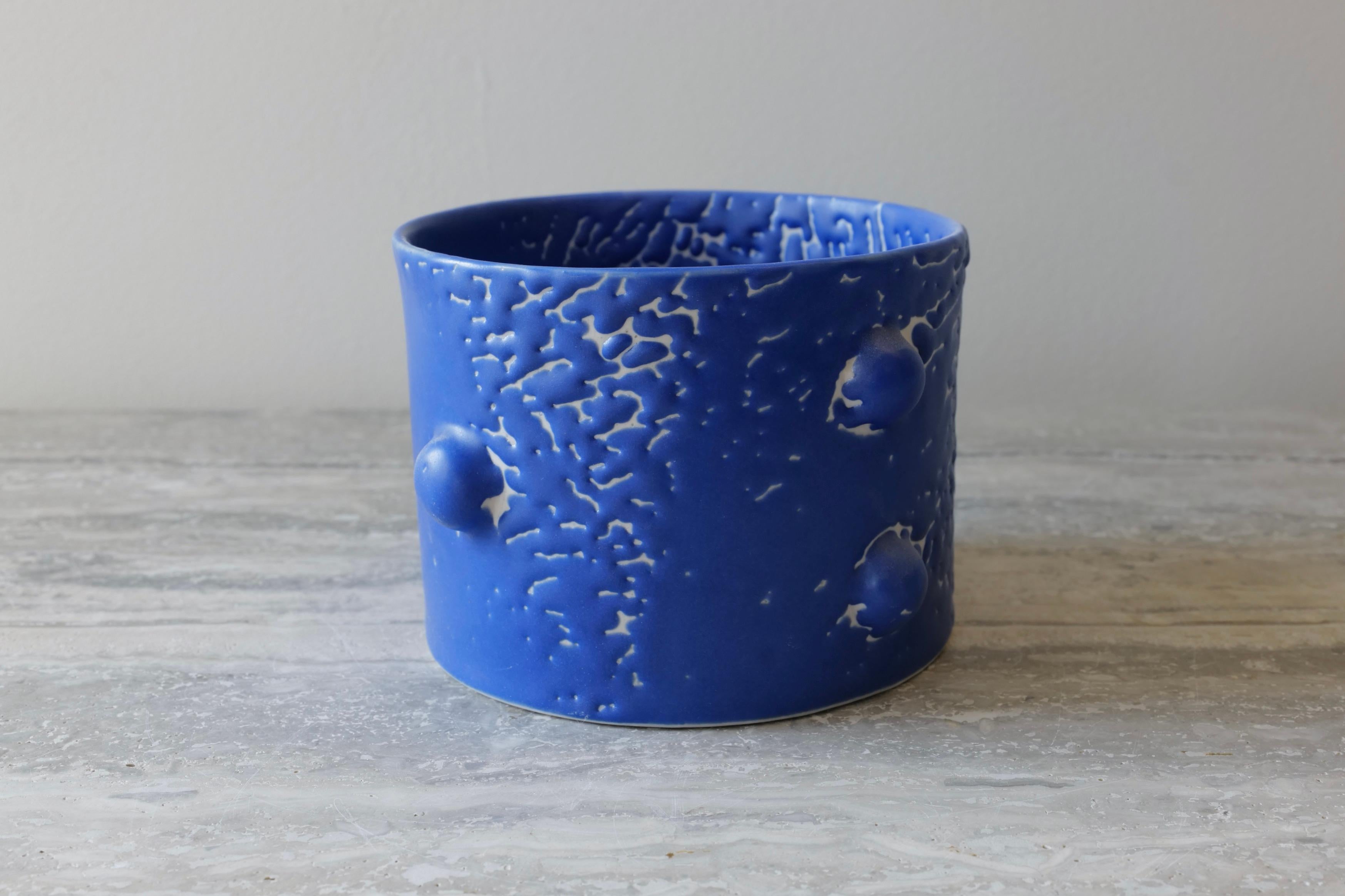 Ce vase court et élégant est moulé à la main en porcelaine et émaillé d'une riche glaçure microcristalline au cobalt qui présente une belle texture craquelée sur toute la surface du récipient. Le motif des 