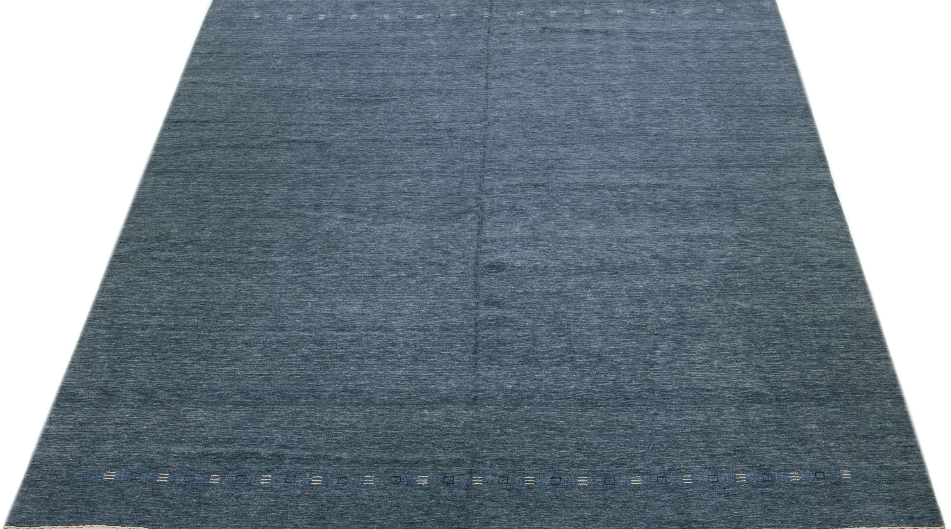This wool rug is handcrafted in the Gabbeh style and features a sophisticated minimalist design. Its dark blue field provides a stunning backdrop for the subtle blue and white accents.

This rug measures 12' x 14'9