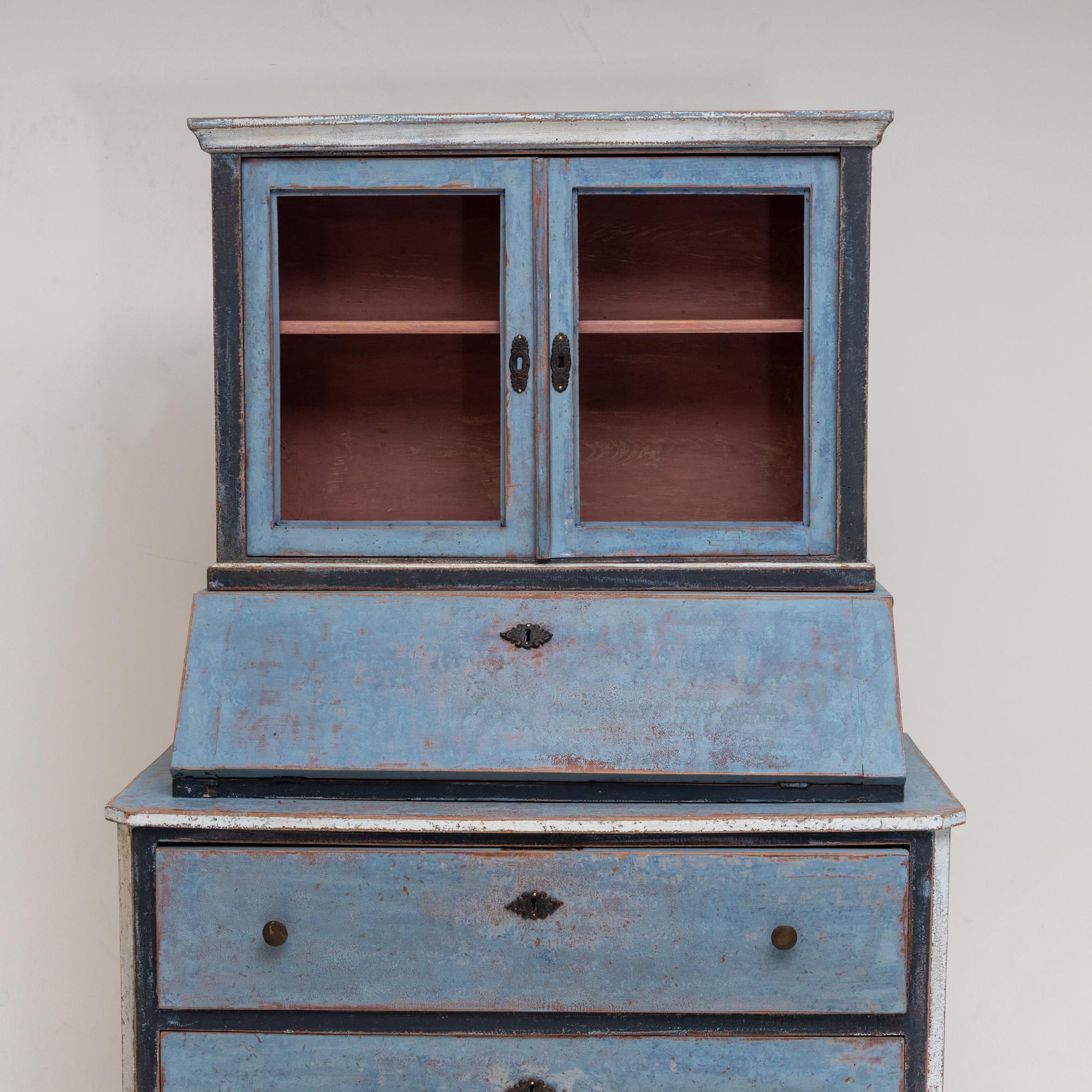 Biedermeier secretary with three-drawer chest of drawers and two-door glazed top and sloping writing flap. The light blue painting is set off in white at the edges and the interior is repainted in brick red. The setting was then given an antique