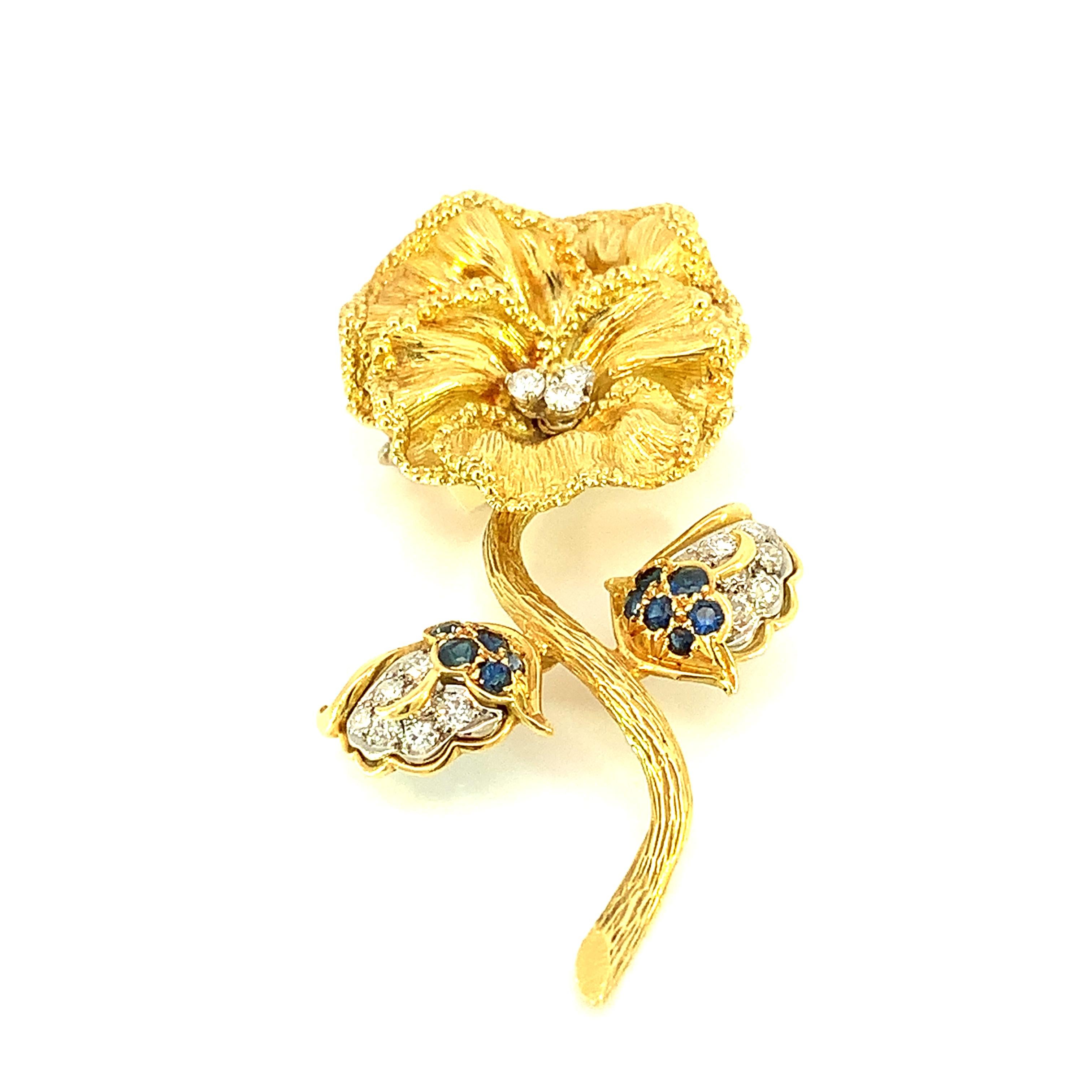 Wir stellen Ihnen unsere exquisite Blumenbrosche mit blauem Saphir und Diamanten vor, ein fein gearbeitetes Schmuckstück, das die zeitlose Eleganz von blauen Saphiren mit dem Glanz von Diamanten vereint, eingefasst in glänzendes 18 Karat Gelbgold.