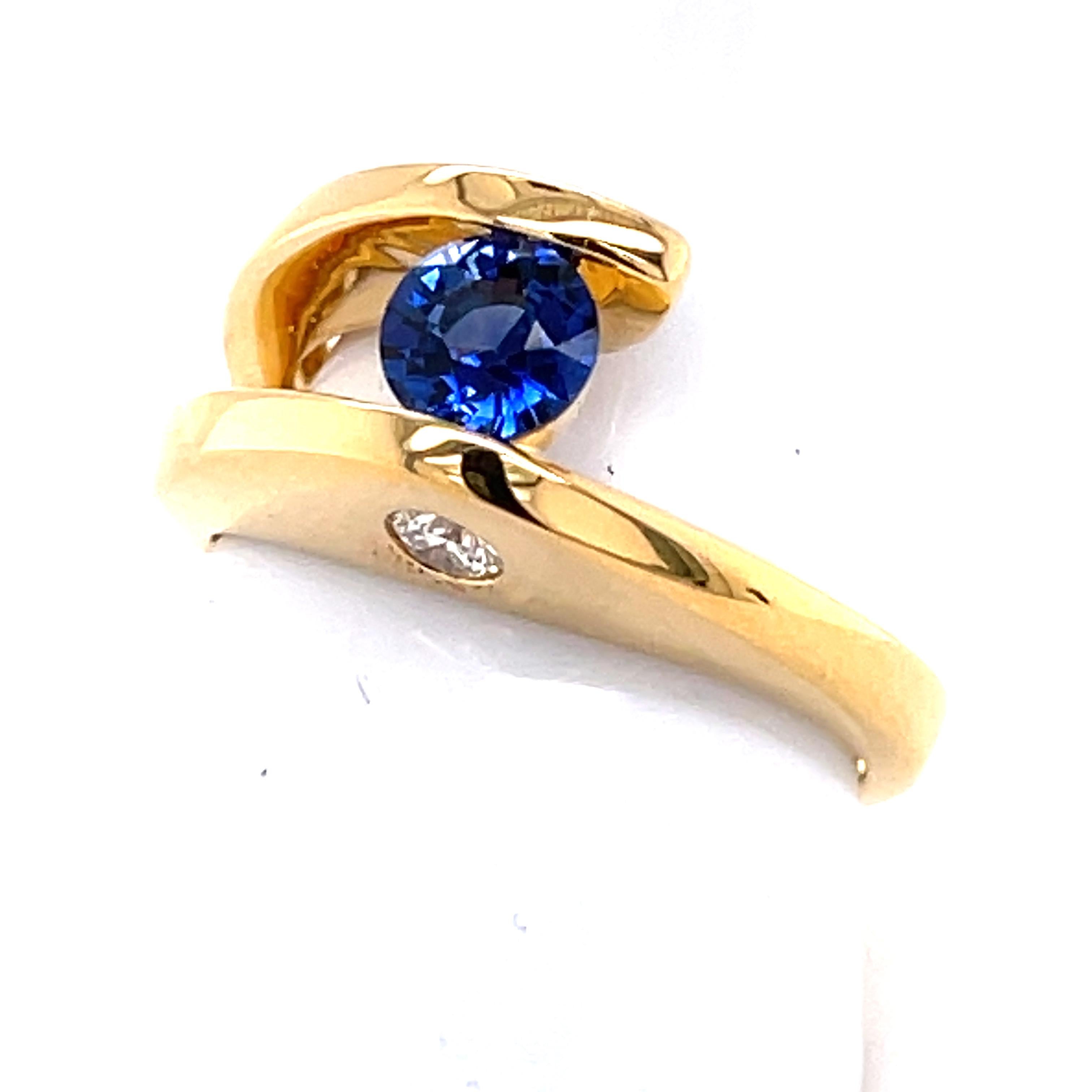 This modern bypass shank ring is done in 18-karat yellow gold. The tension set blue Sapphire center stone has a small basket for extra security  The one VS clarity and G color diamond is a surprise on the side of the ring and can be shown off to