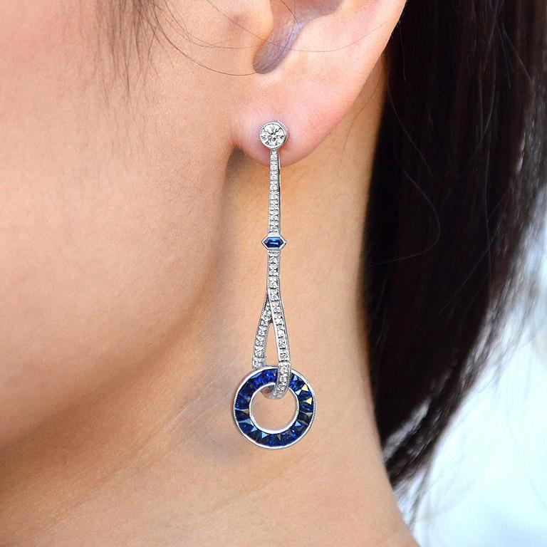 These fabulous Art Deco style blue sapphire and diamond drop earrings feature French cut channel set sapphires in milgrain edged 18k white gold circlet. The earrings are hung from the straight bars of approximately 0.80 carat diamonds adorned with a
